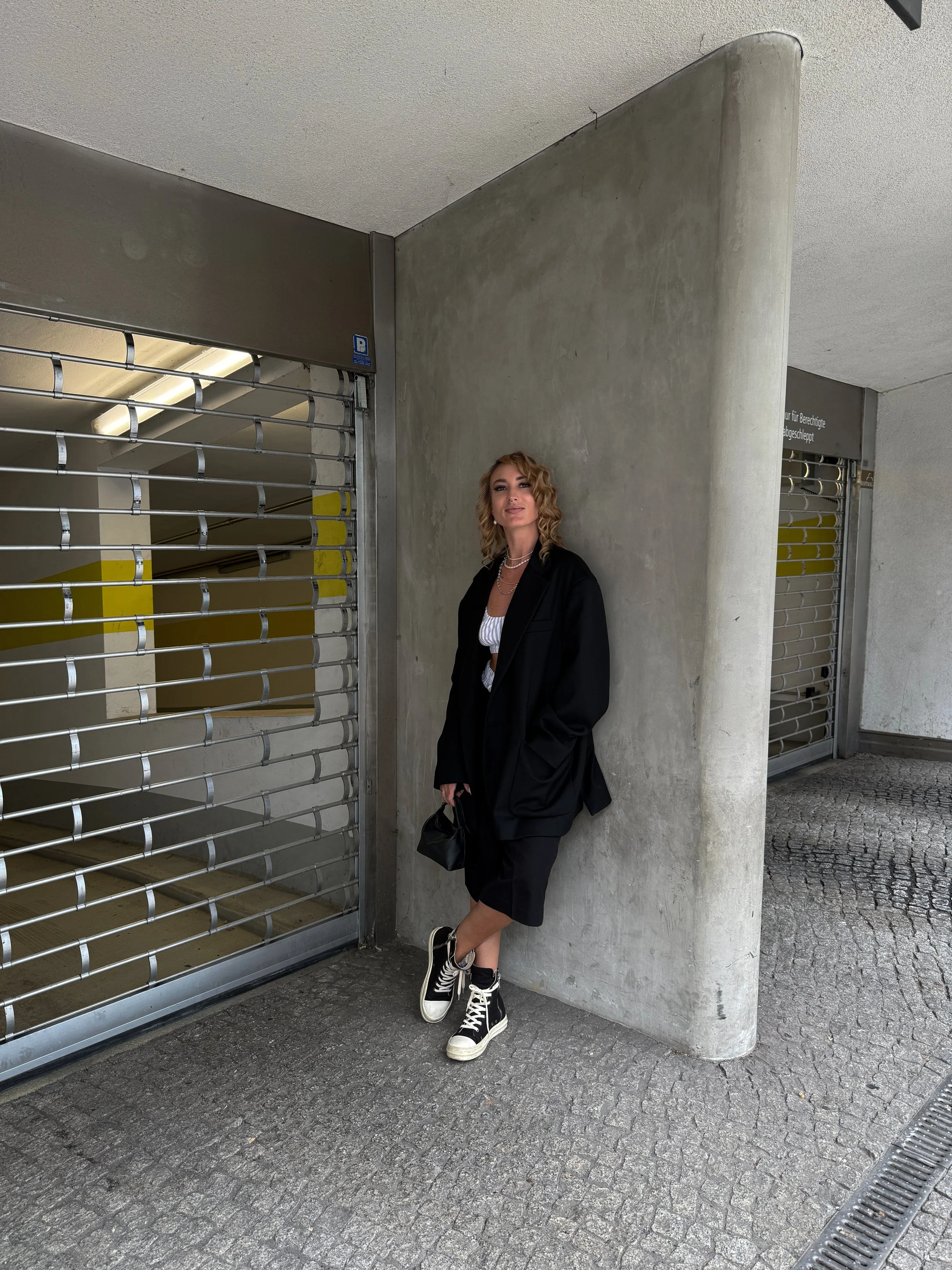 Junge Frau in schwarzer Jacke, weißen Turnschuhen und Minikleid, die vor einer grauen Betonwand an einer Rollgittertür steht, während sie die Kamera anlächelt.
