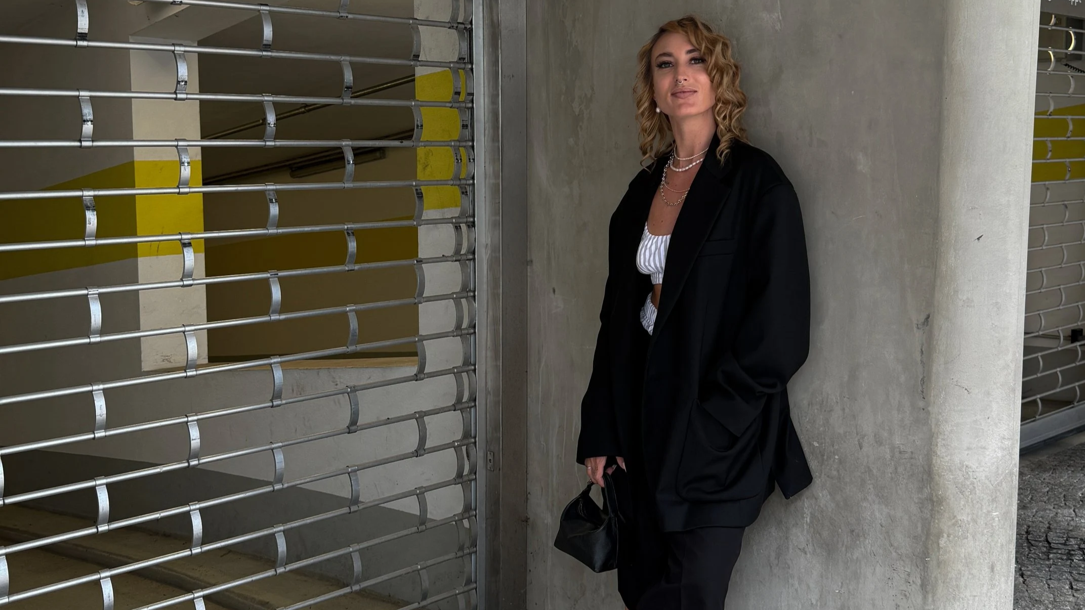 Eine Frau mit lockigem, blondem Haar, bekleidet mit einer schwarzen Jacke, weißen Top und mehreren Perlenketten, lehnt an einer Betonsäule neben einem Metallrolltor in einer modernen Parkgarage.