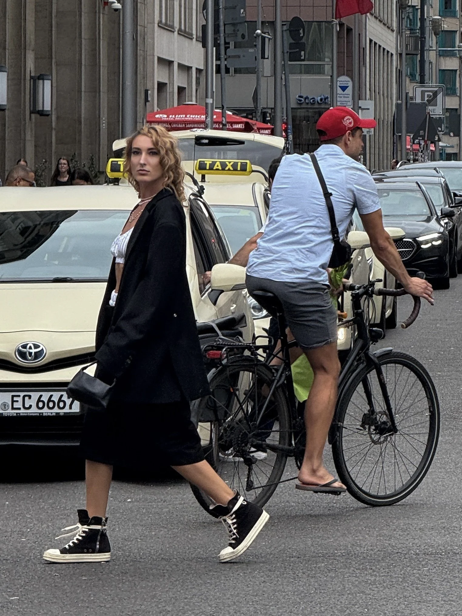 Ein Mann auf einem Fahrrad und eine weibliche Fußgängerin, beide in der Innenstadt, mit Autos und Gebäuden im Hintergrund.