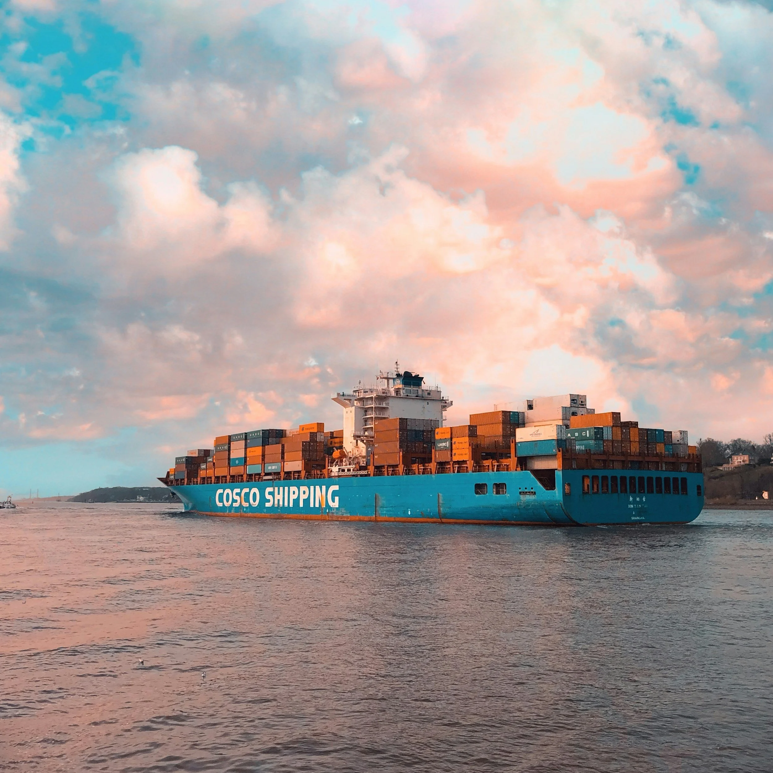 A large cargo ship carrying numerous shipping containers is sailing on a body of water during sunset or sunrise, with a partly cloudy sky and distant land visible in the background.