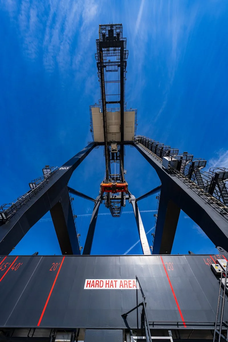 Looking up at a tall construction crane against a blue sky, with a safety warning sign that reads 'HARD HAT AREA' at the base.