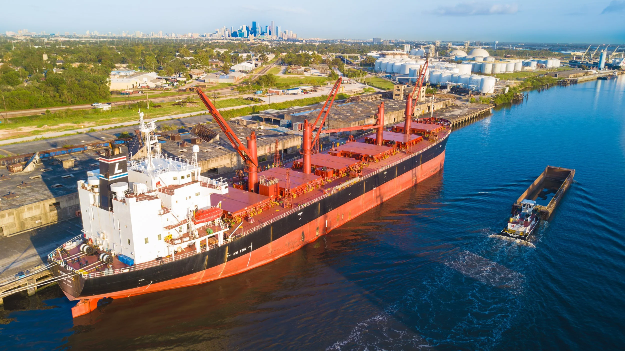 Aerial view of a large red and black cargo ship docked at a port with storage tanks and factory buildings nearby, and a city skyline in the background.