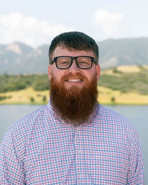 A smiling man with glasses and a full beard outdoors, standing in front of a lake and green hills.