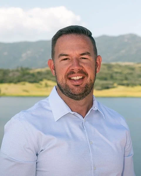 A man with short dark hair and a beard smiling outdoors near a body of water with hills in the background, wearing a white dress shirt.