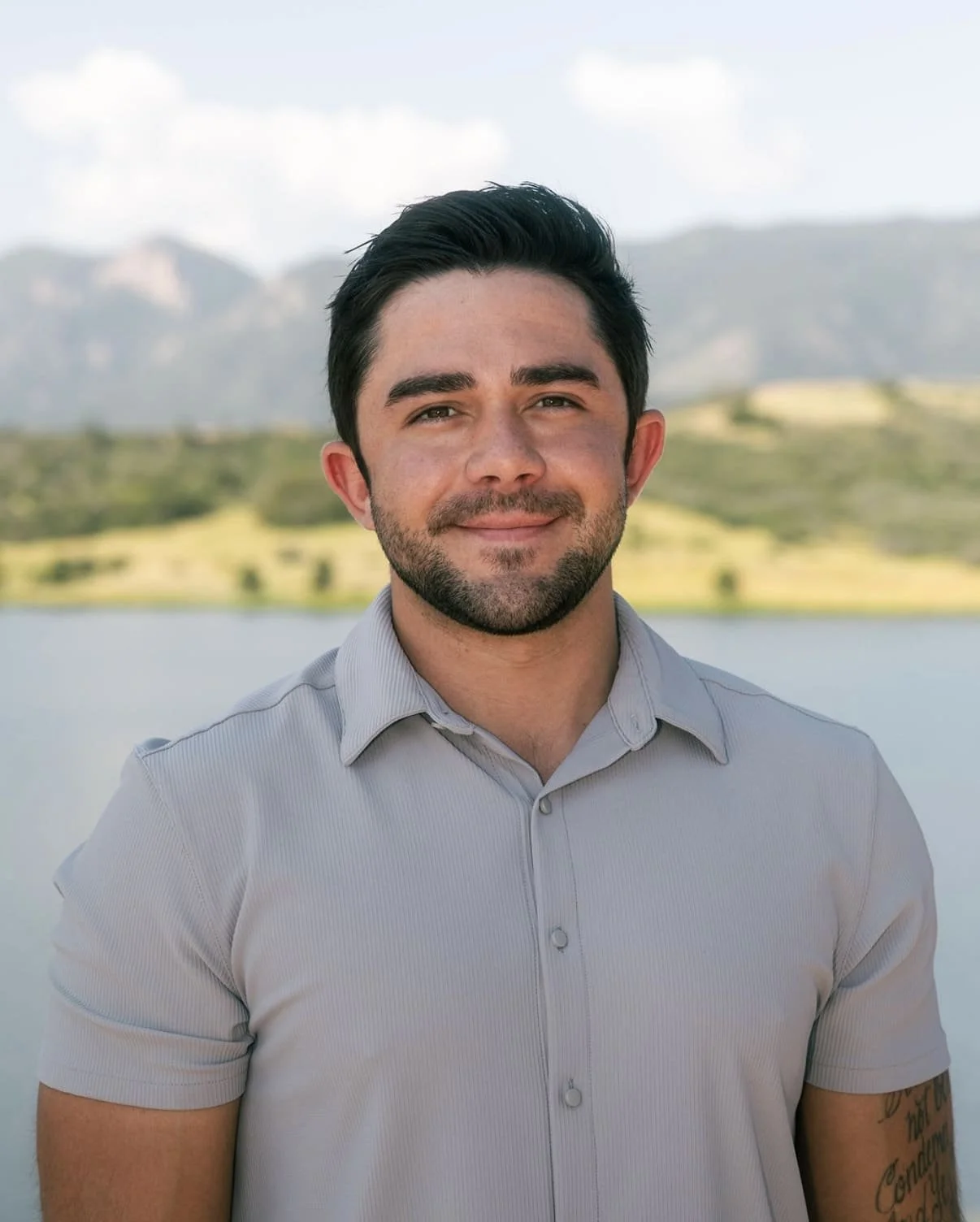 A young man with dark hair and a beard, smiling, standing outdoors near a body of water with mountains and hills in the background, wearing a light gray short-sleeved button-up shirt.