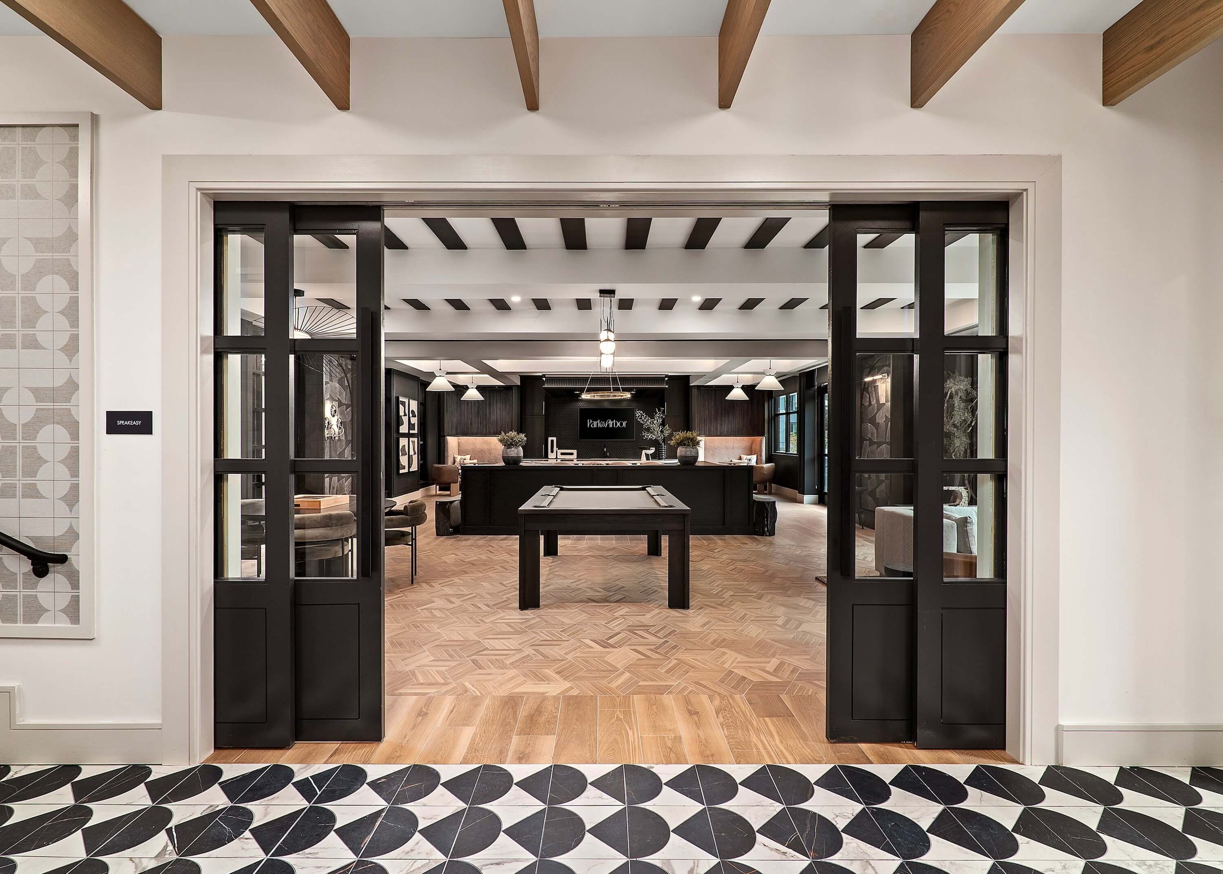 View of an interior space with a reception desk in the distance, black decorative wooden panels, pendant lights, and black and white patterned flooring.