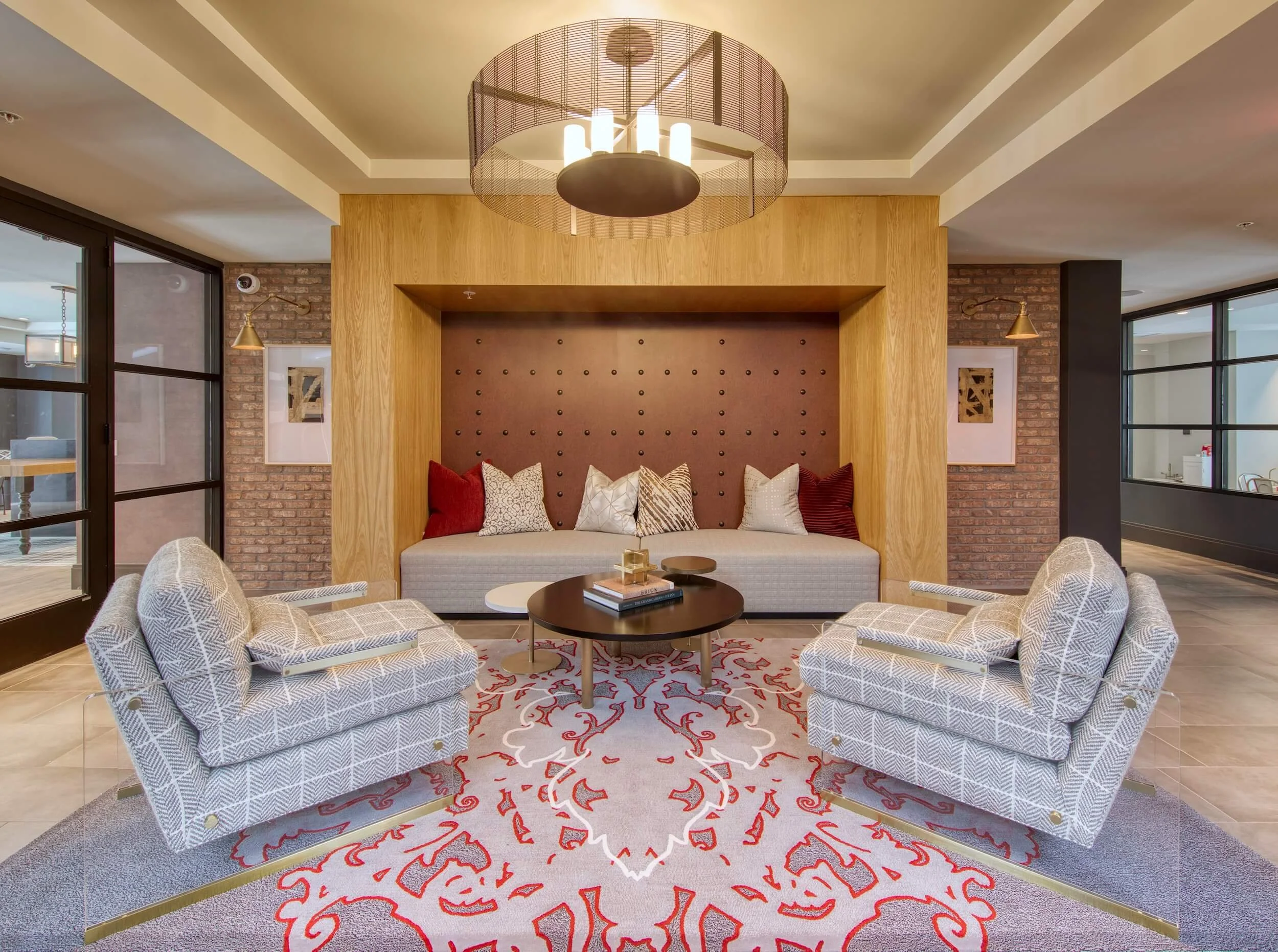 A modern hangout space with a wall nook featuring cushions, surrounded by brick and wood wall accents, with large windows, and a chandelier overhead.