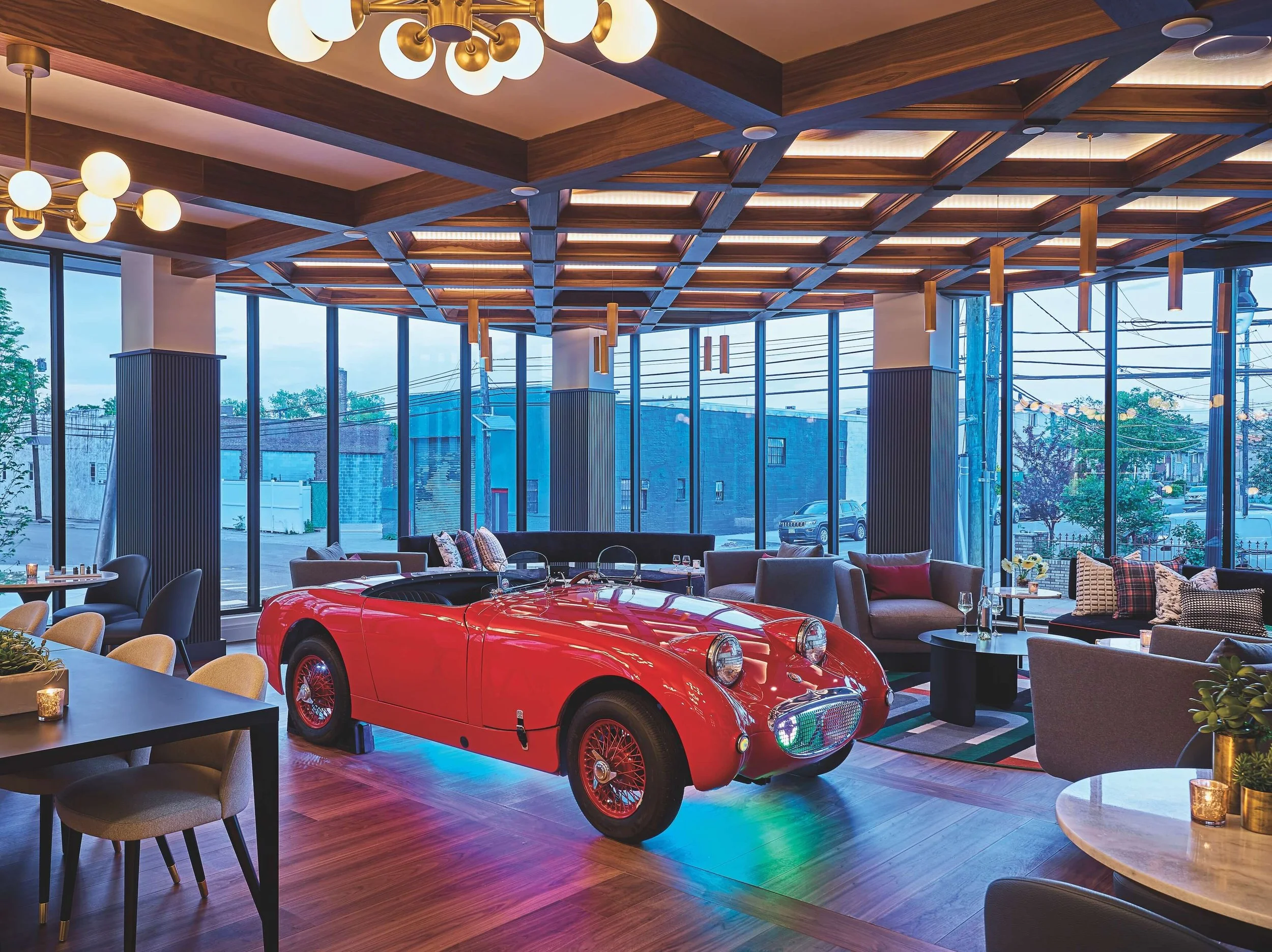 Lounge with large windows, wooden ceiling, and stylish lighting. A red vintage car is parked inside as a decorative element.