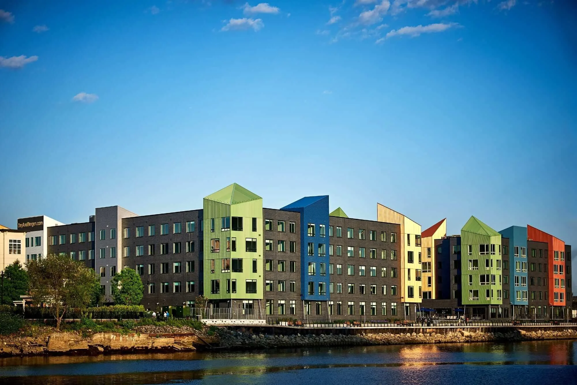 Colorful modern apartment building facing a waterfront with trees in the foreground and a blue sky with a few clouds.