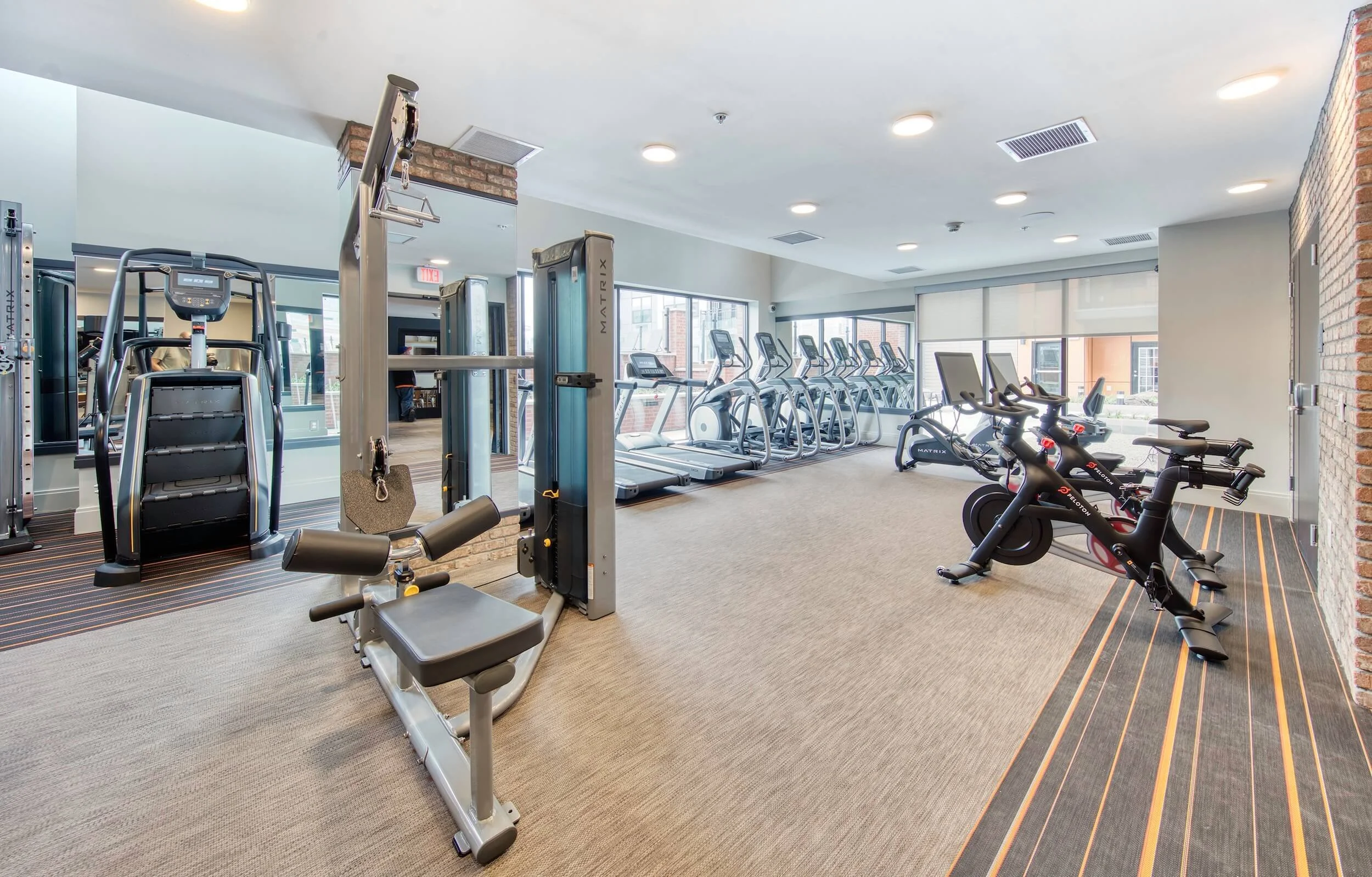 A fitness center with various exercise equipment including treadmills, stationary bikes, and strength training machines, with large windows letting in natural light.