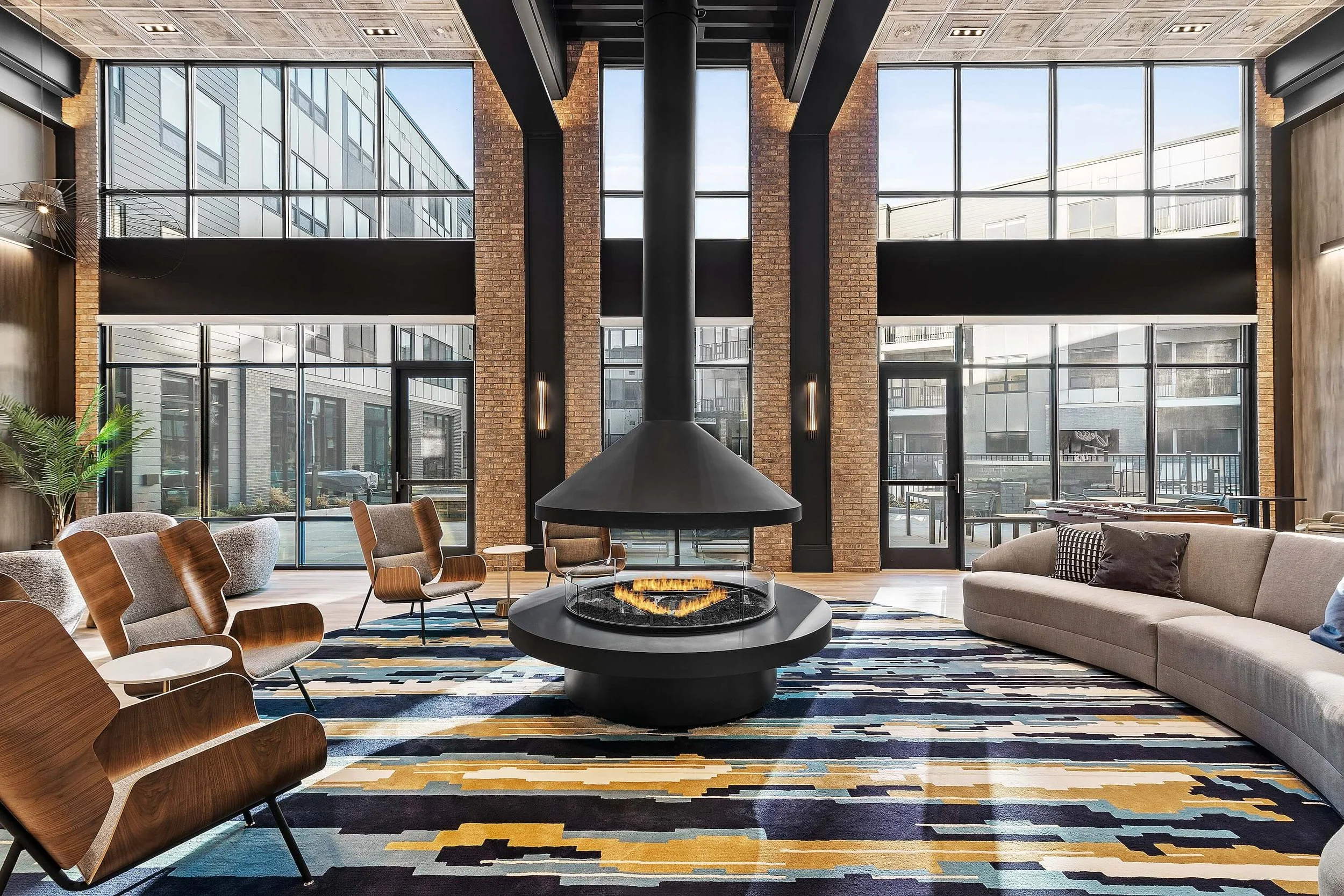 Modern lobby with large floor-to-ceiling windows, a central black fireplace, and seating including a beige sofa and wooden chairs, with a colorful geometric-patterned rug.