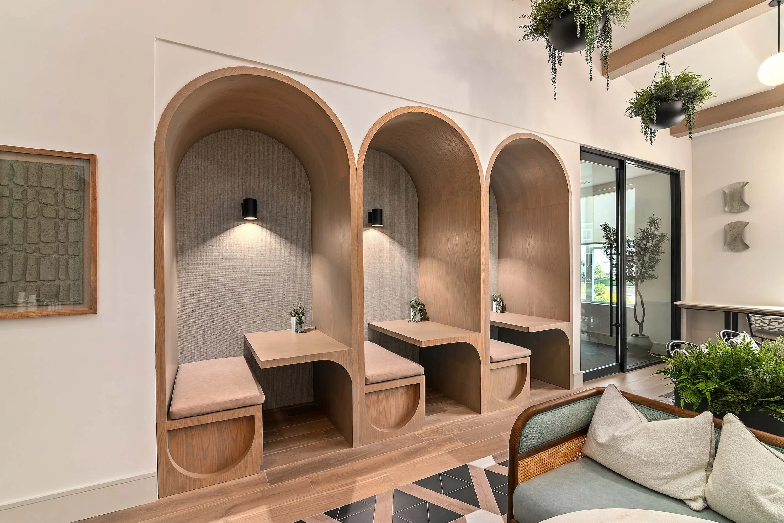 Modern interior with decorative wooden arches used as seating or partitions, small potted plants on each section, hanging greenery arrangements from the ceiling, a sliding glass door, and minimalist wall art.