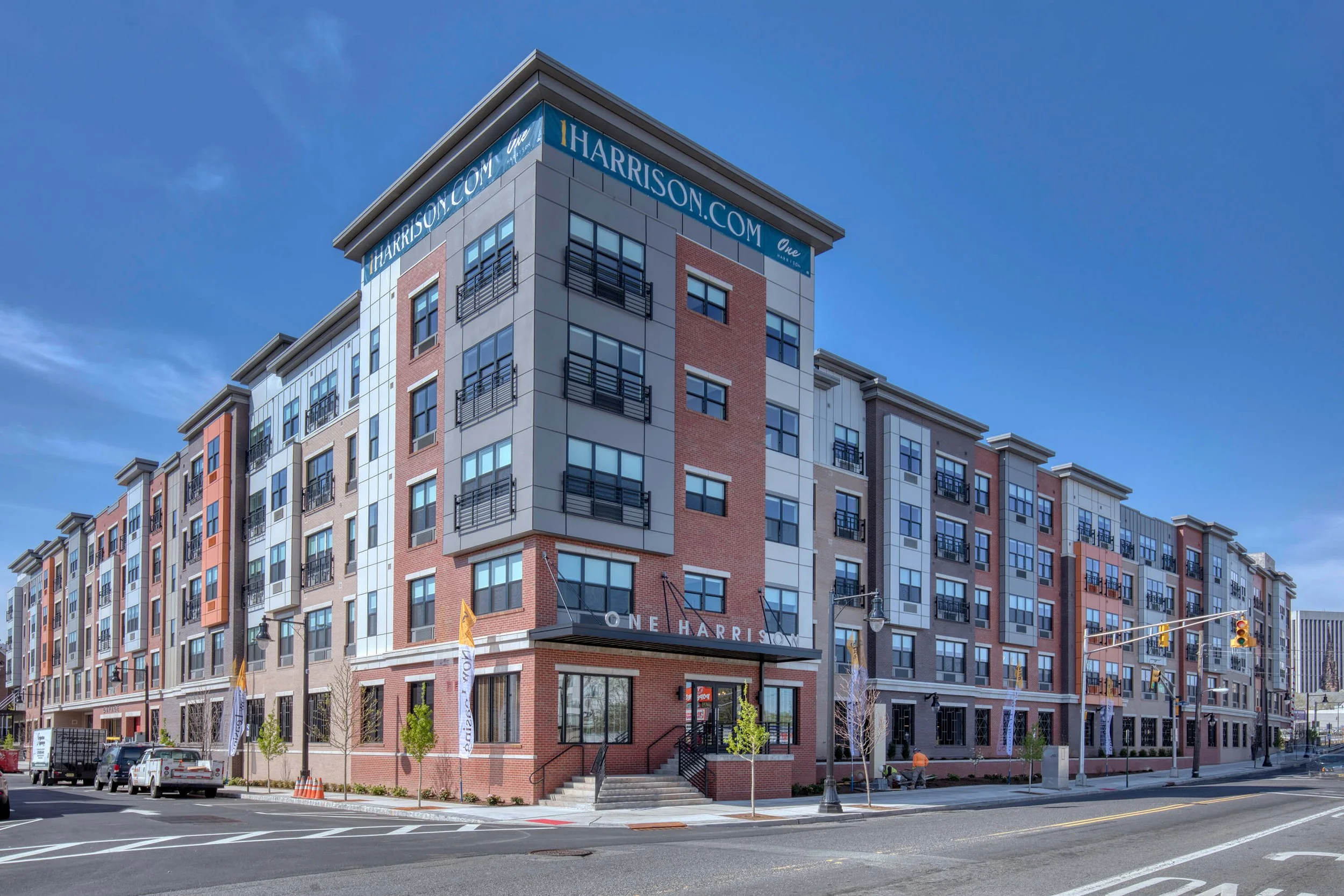 Modern multi-story apartment building with colorful facade and signs displaying 'harrison.com' and 'One Harrison' at corner, with parked cars, small trees, and traffic lights in foreground.