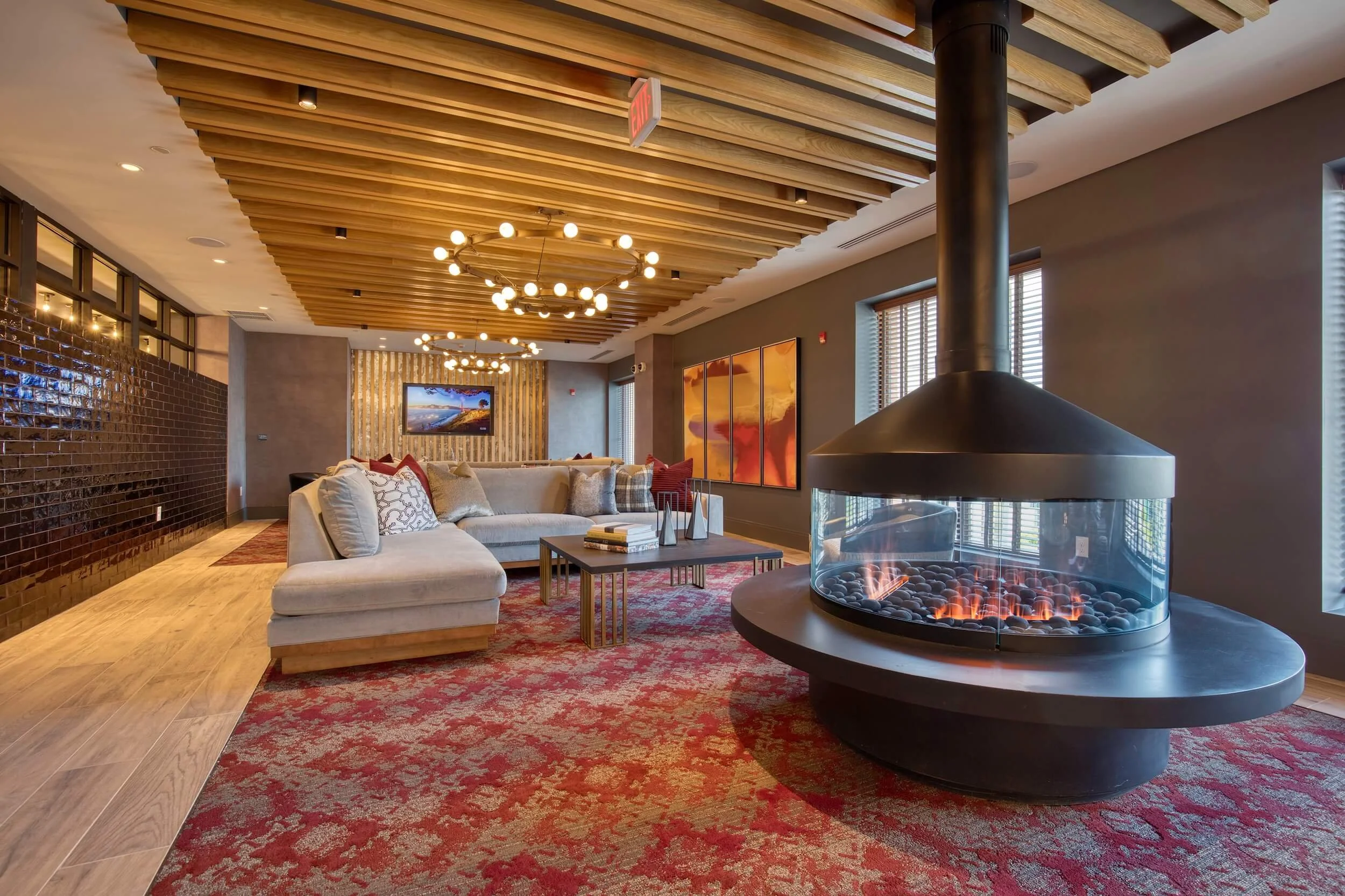 A modern lobby with a wooden slatted ceiling, a chandelier with globe lights, a fireplace with transparent glass, a large beige sofa with pillows, artwork on the walls, windows with blinds, and a red patterned rug.