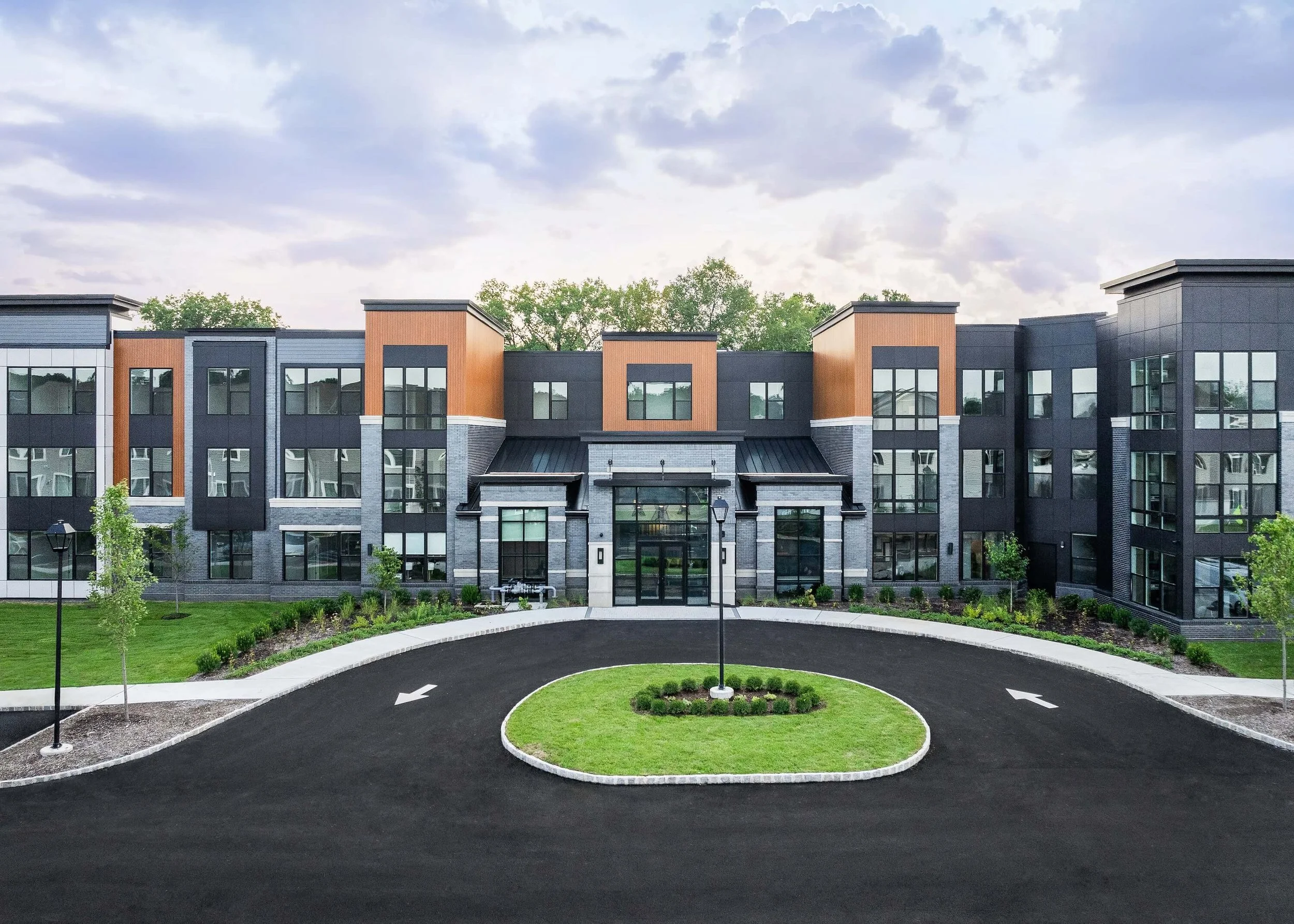 Modern multi-story residential buildings with colorful brick and black accents, surrounded by a landscaped yard, small trees, and lamp posts under a partly cloudy sky.