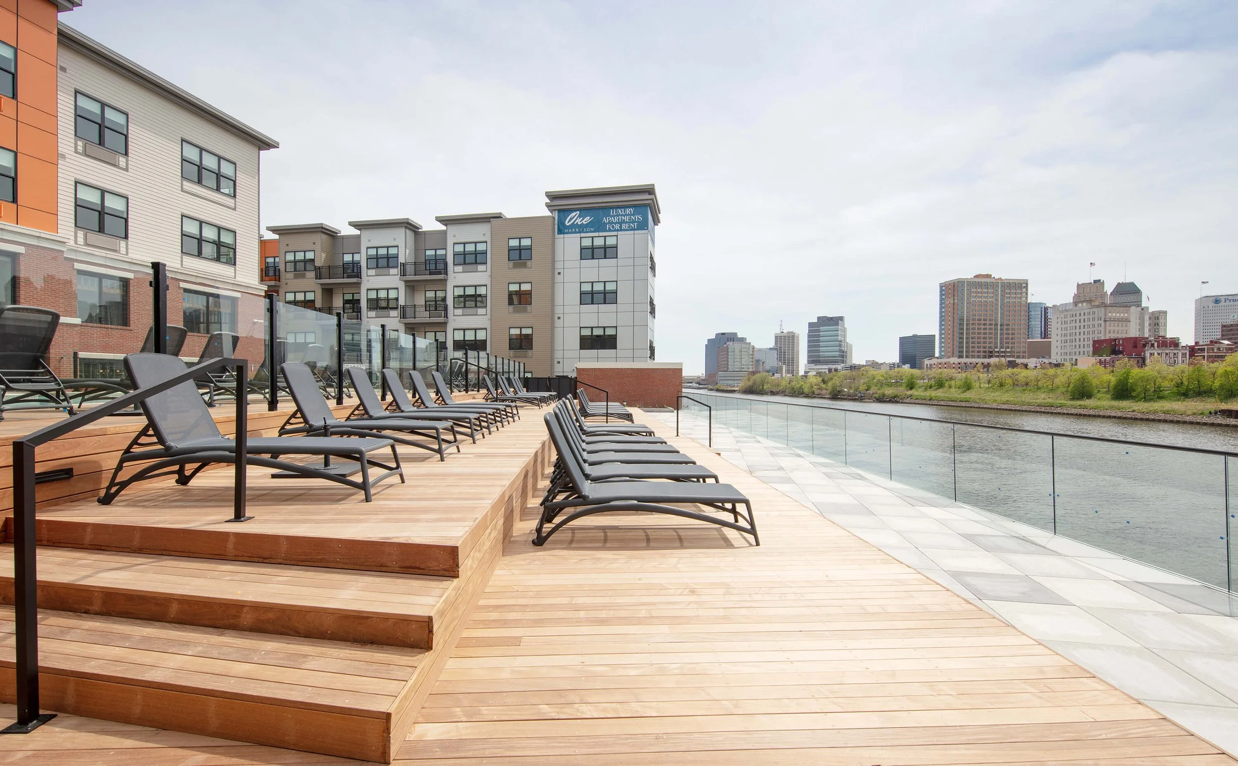 Outdoor rooftop deck with black lounge chairs, glass railing, overlooking city skyline.