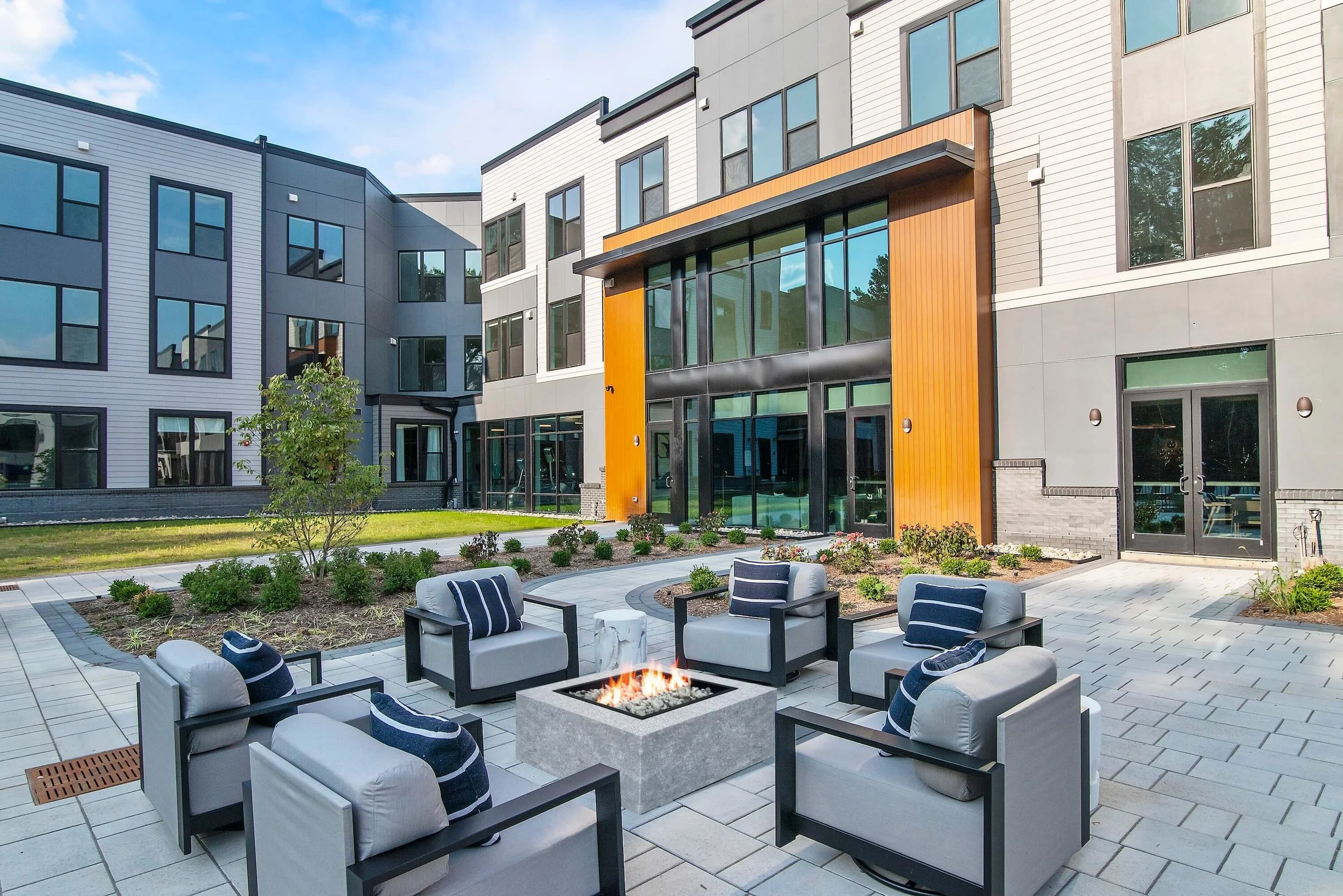 Modern apartment building with a courtyard featuring patio chairs arranged around a fire pit, and a small landscaped area with plants and a tree, under a partly cloudy sky.