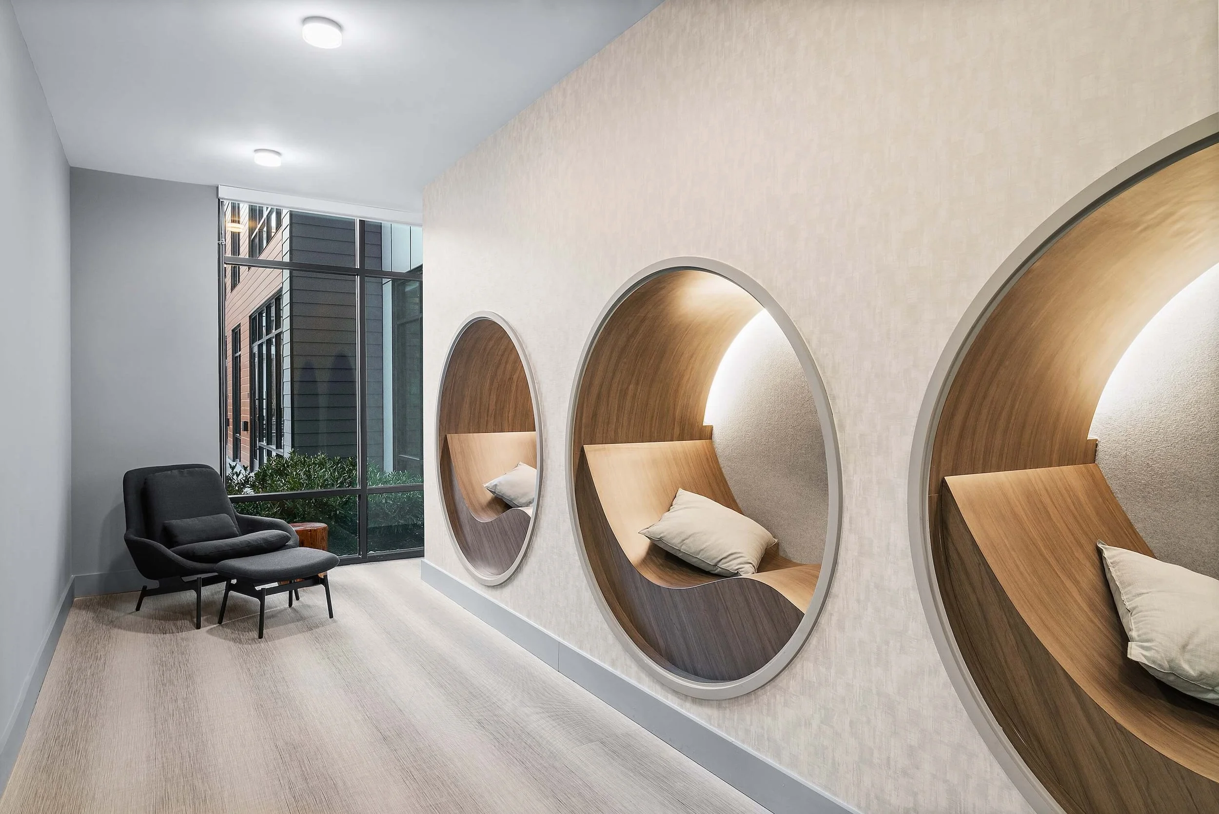 A modern interior space with a wall featuring circular wooden seating sections each with a pillow. There is a black armchair and a small side table next to a large window showing an exterior building with dark siding.