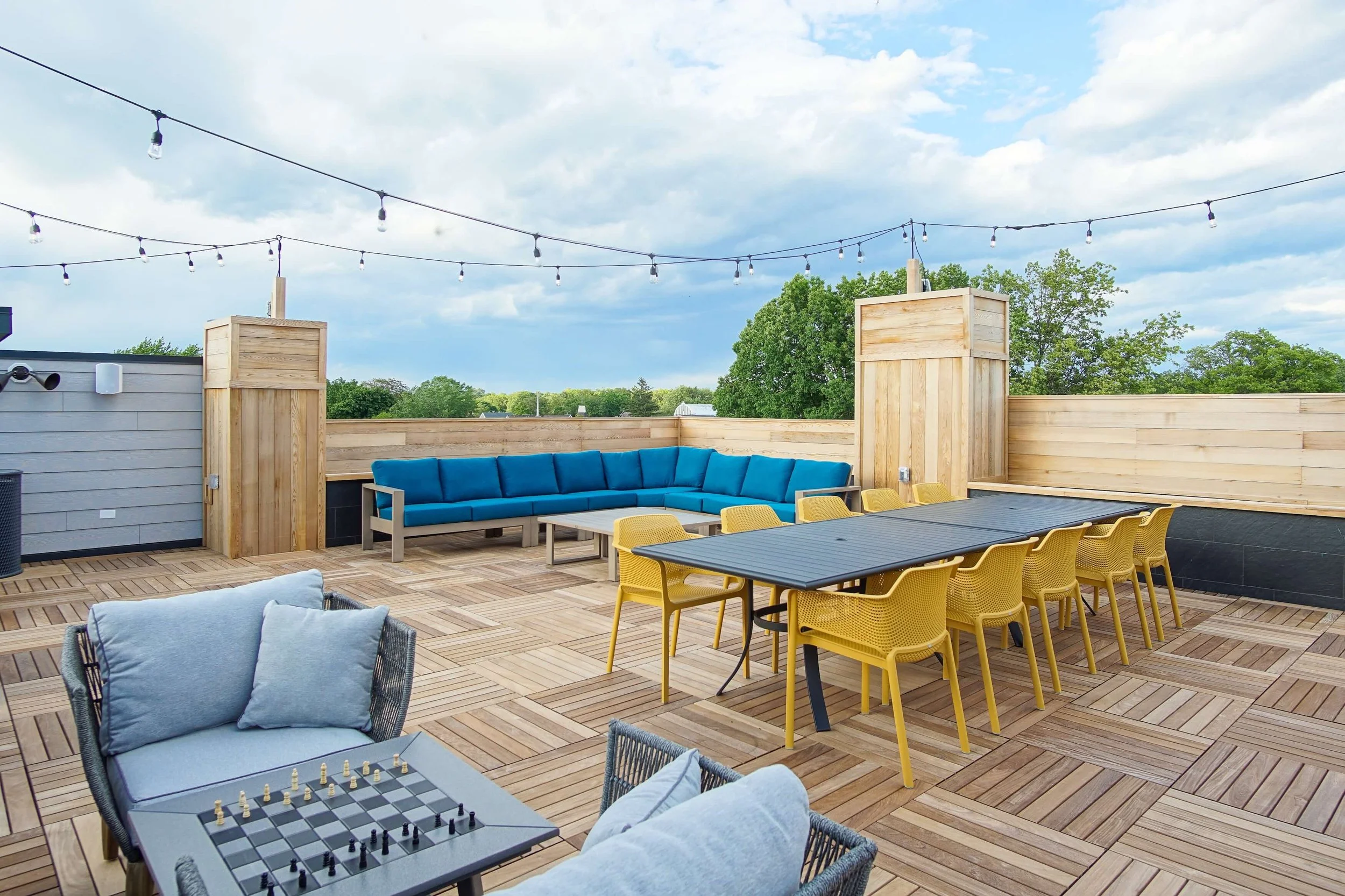Outdoor rooftop patio with wooden deck, yellow chairs, blue outdoor couch, chessboard table, string lights, and a view of green trees and cloudy sky.