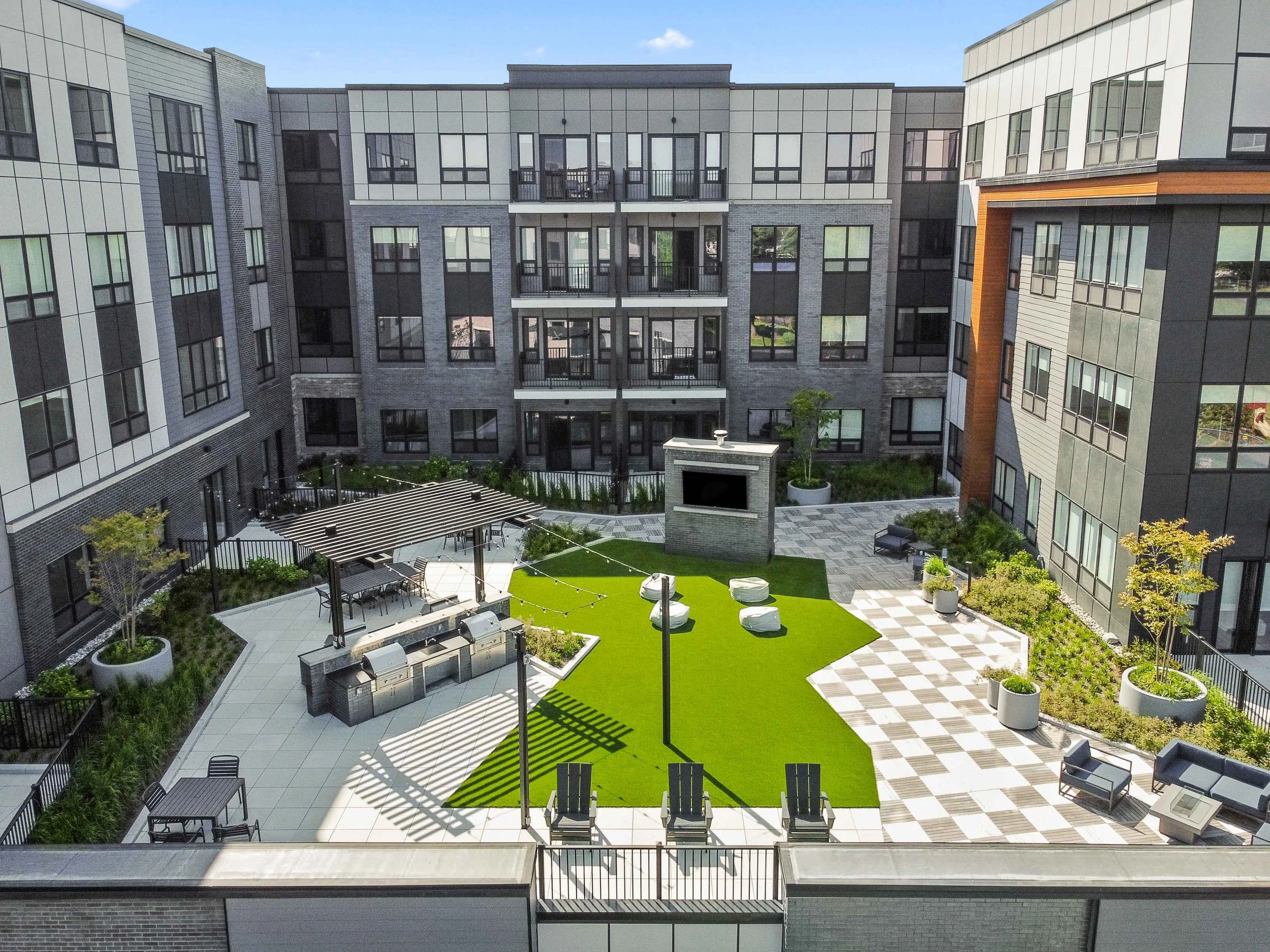A modern apartment complex courtyard with outdoor seating, a grassy area with white lounge chairs, trees, string lights, and a fire pit with a building in the background.