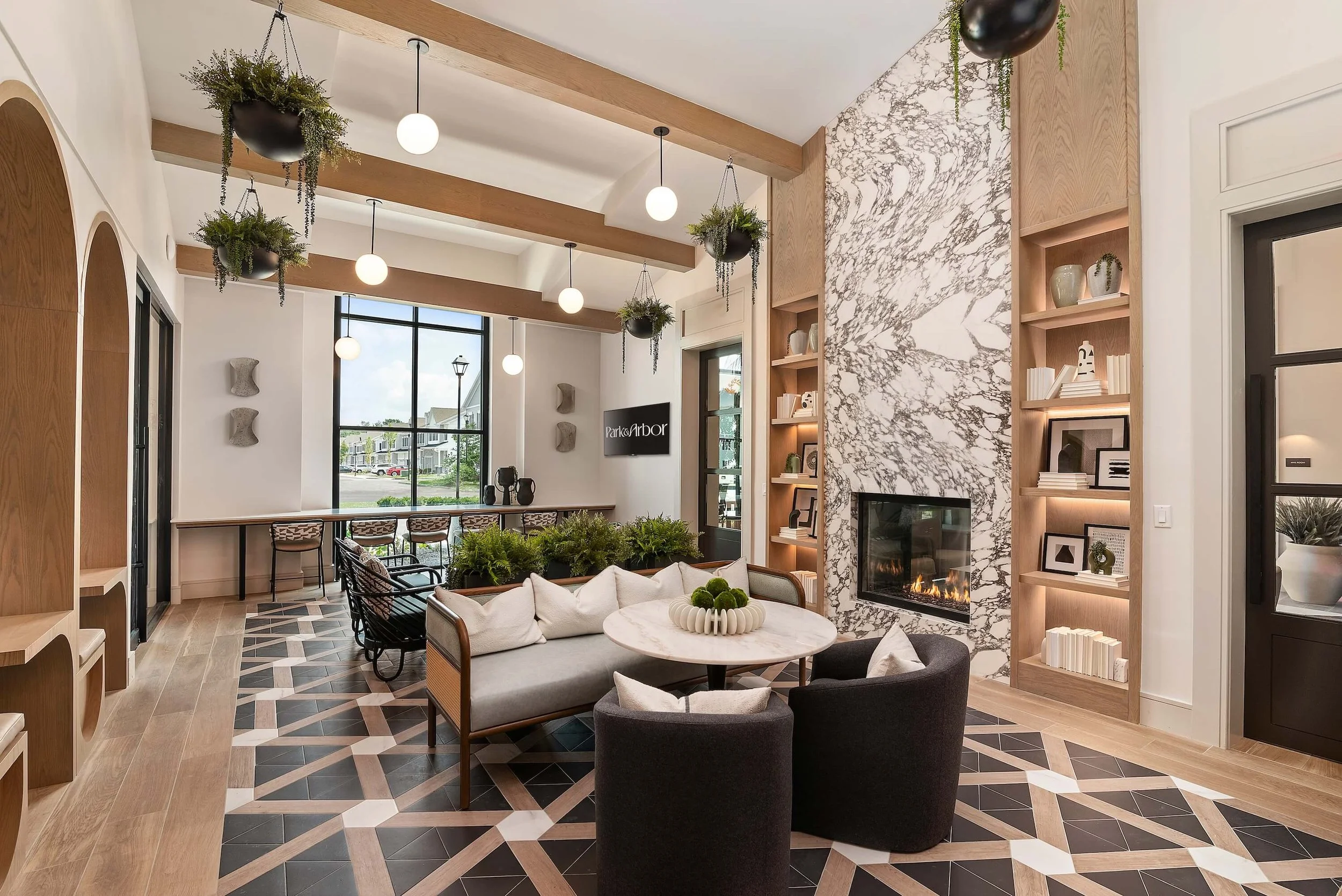 Modern lobby with wood beams, hanging plants, round pendant lights, a marble fireplace, built-in shelves with decor, and seating with cushions. Large window showing a street view.