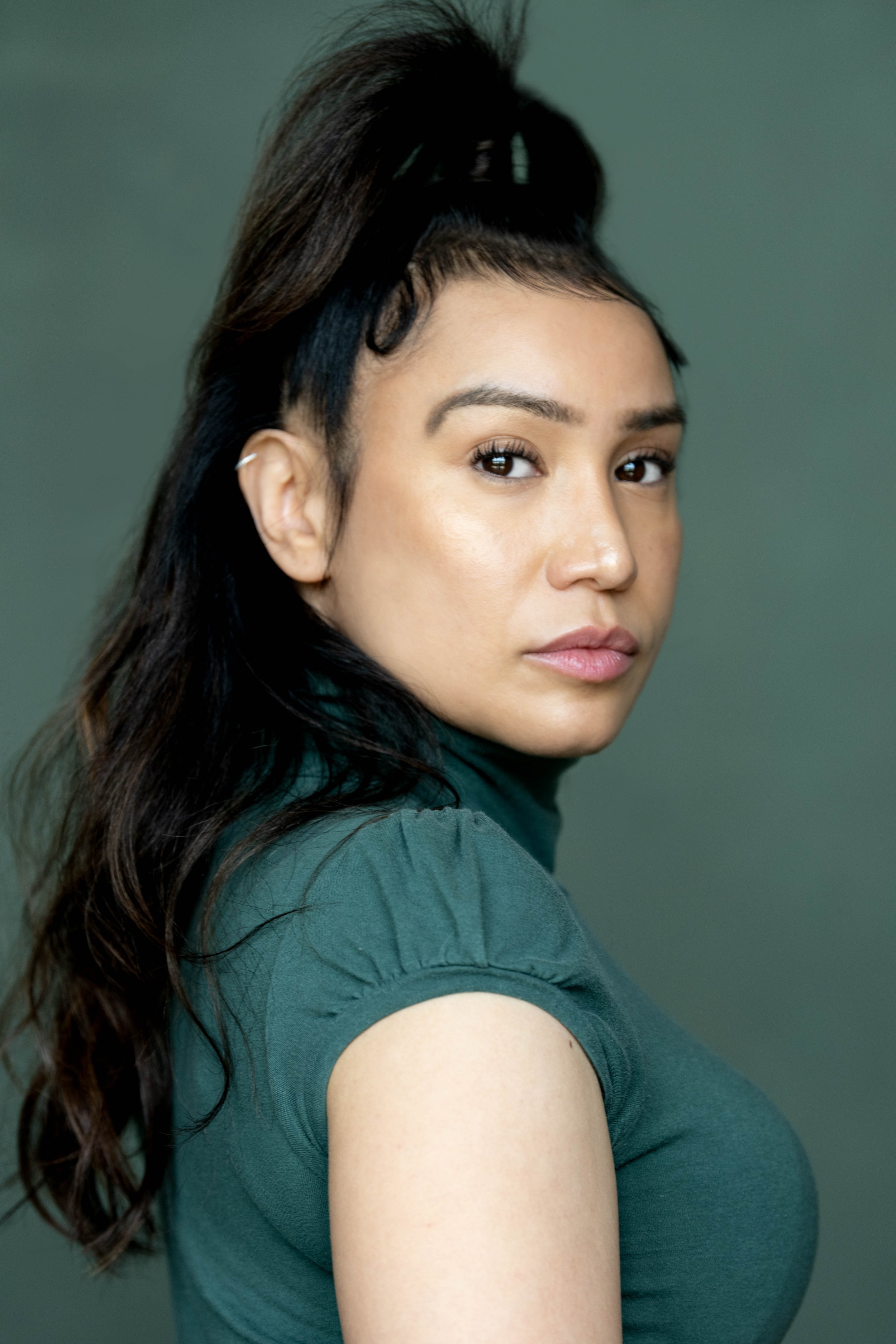 Close-up of a woman with dark hair styled in an elaborate updo, wearing a dark green top and a small earring, against a muted green background.