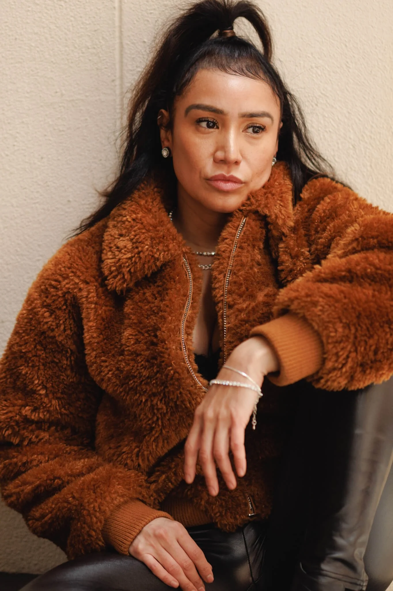 A woman with dark hair styled in a high ponytail, wearing a brown fuzzy jacket, jewelry, and black leather pants, sitting against a beige wall.