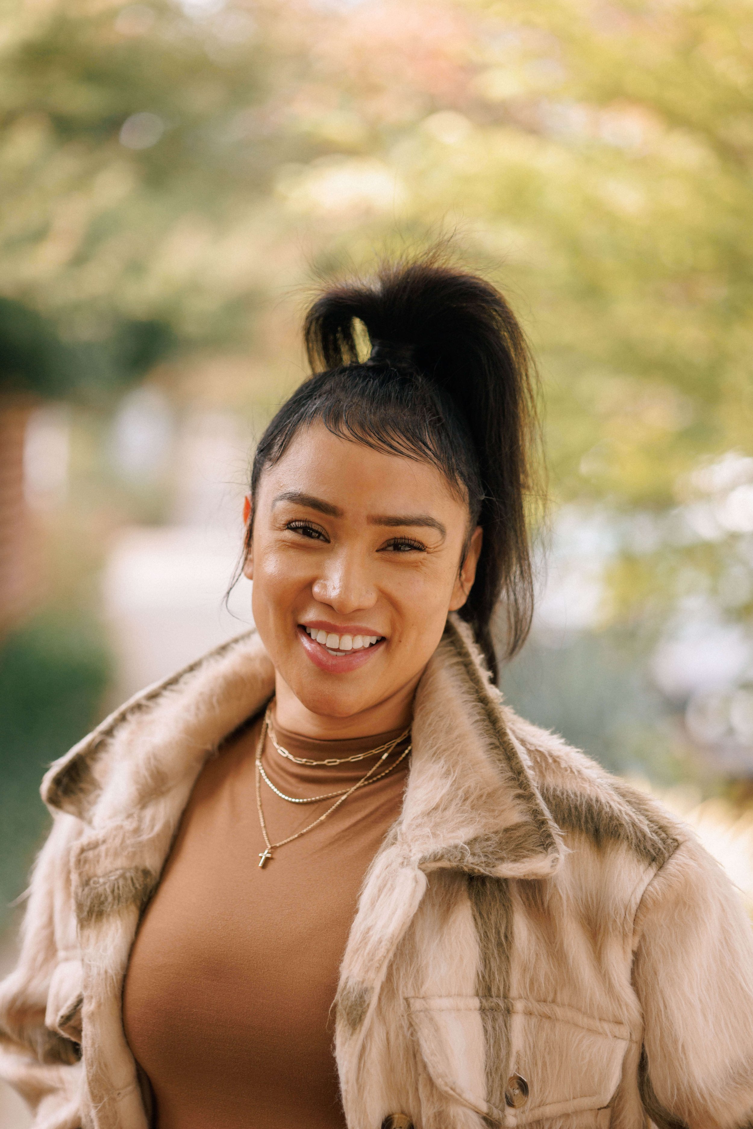 A young woman with a high ponytail smiling outdoors, wearing a tan shirt, layered necklaces, and a fur-lined beige jacket with a blurred autumn background.