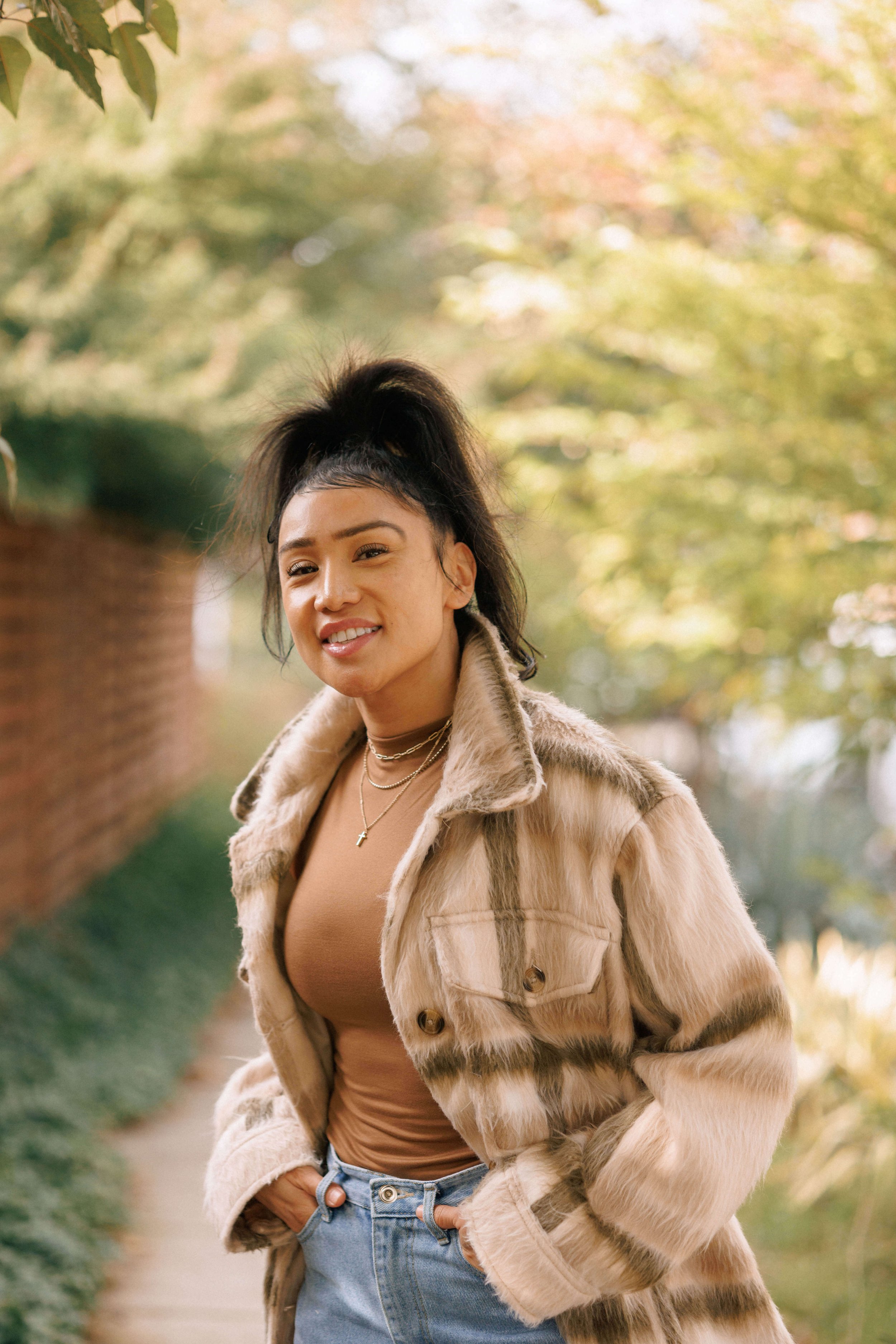 A young woman walking outdoors in fall foliage, wearing a beige and brown fur coat, a tan top, and blue jeans, with her hands in her pockets and smiling.
