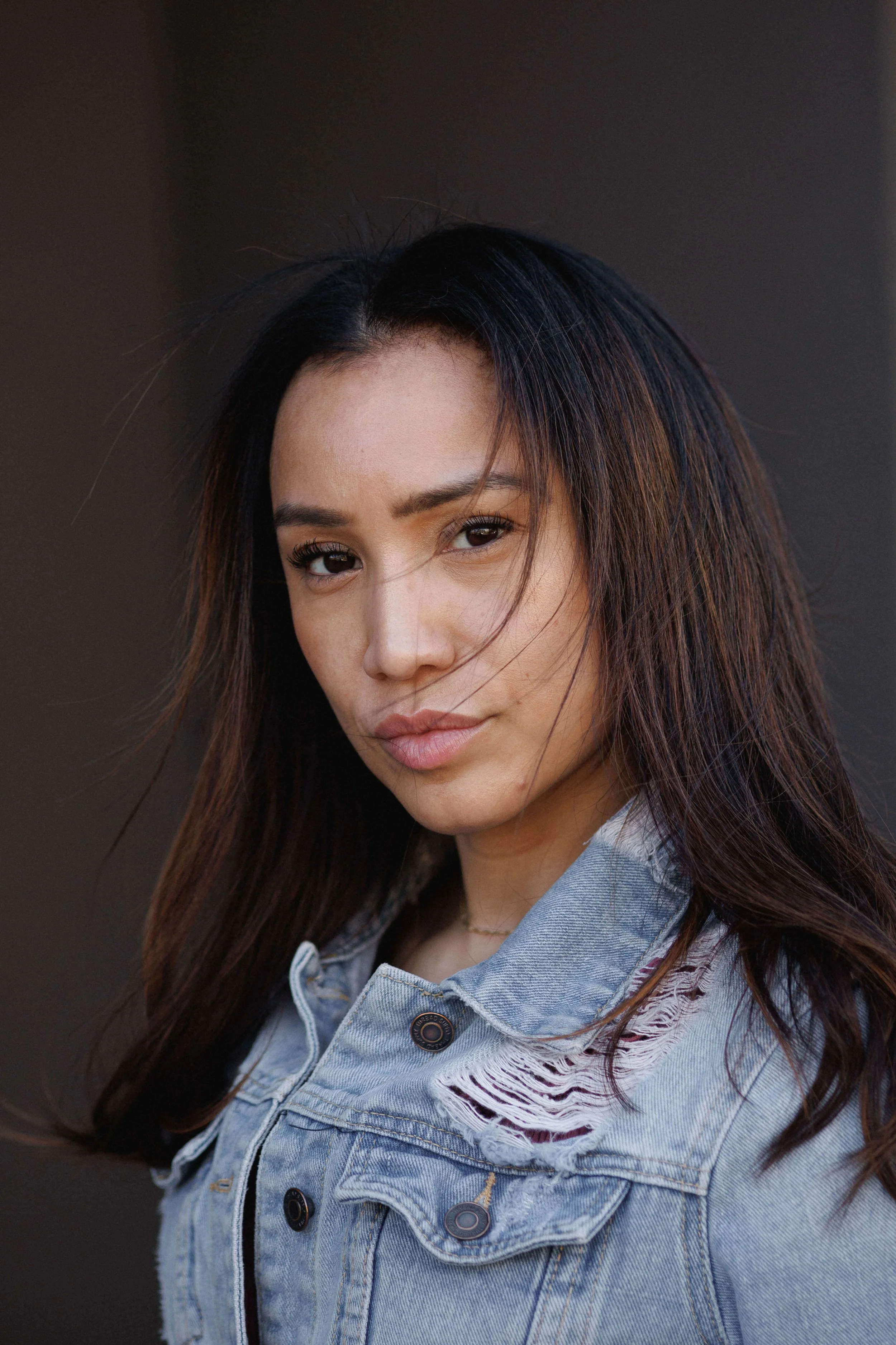 A young woman with long dark hair, wearing a light blue denim jacket with frayed edges and buttons, looking directly at the camera against a dark background.