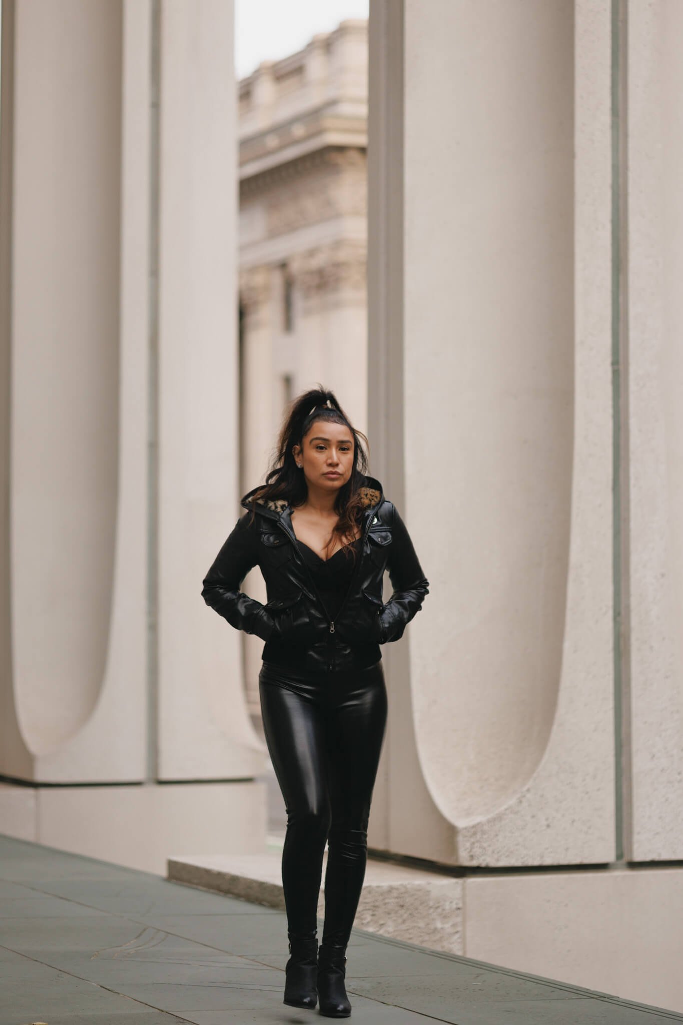 A woman with dark hair in a high ponytail, wearing a black leather jacket and shiny black pants, standing in front of modern architectural structures with curved white walls.