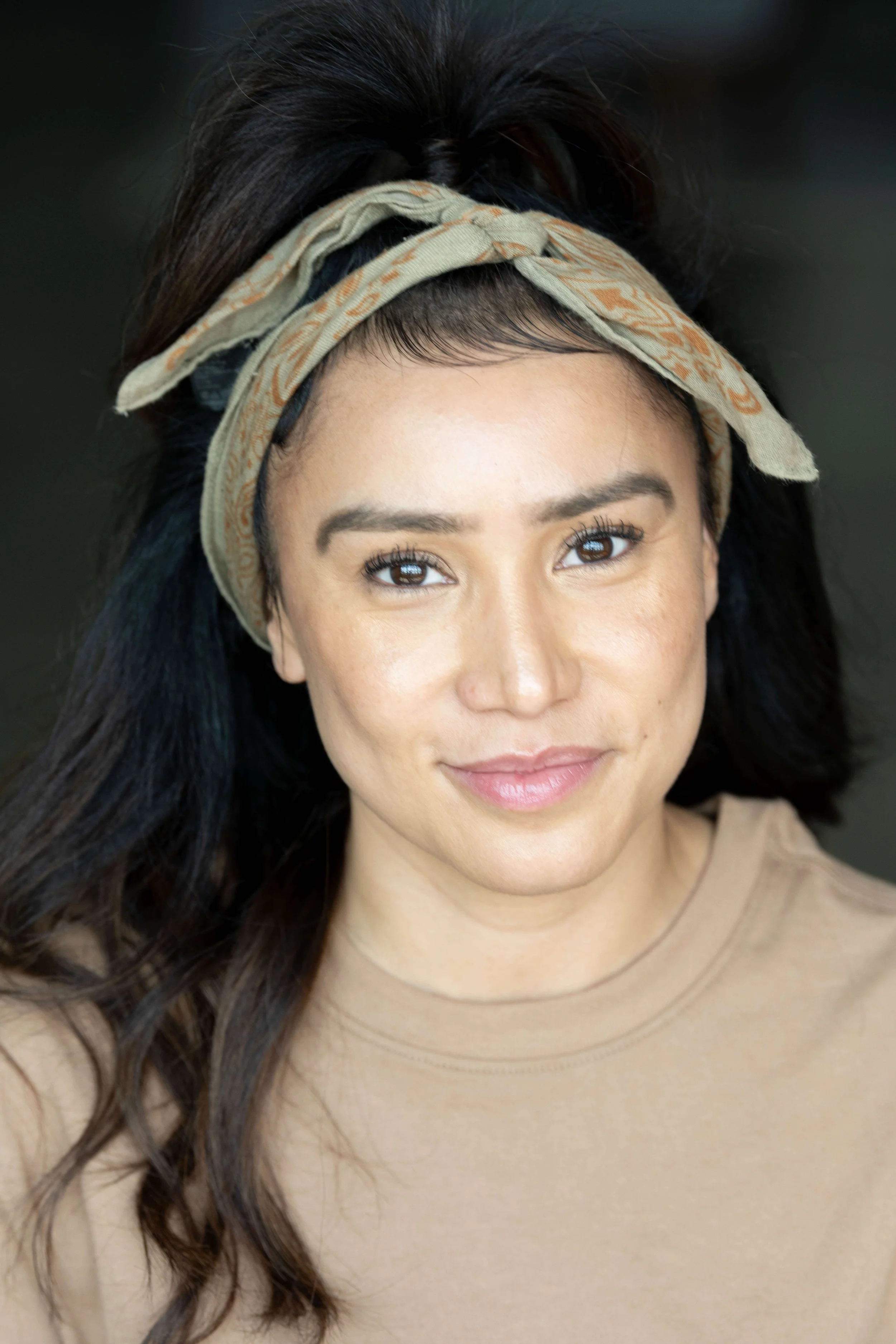 Close-up of a woman with long dark wavy hair, wearing a beige top and a patterned headband, smiling softly.