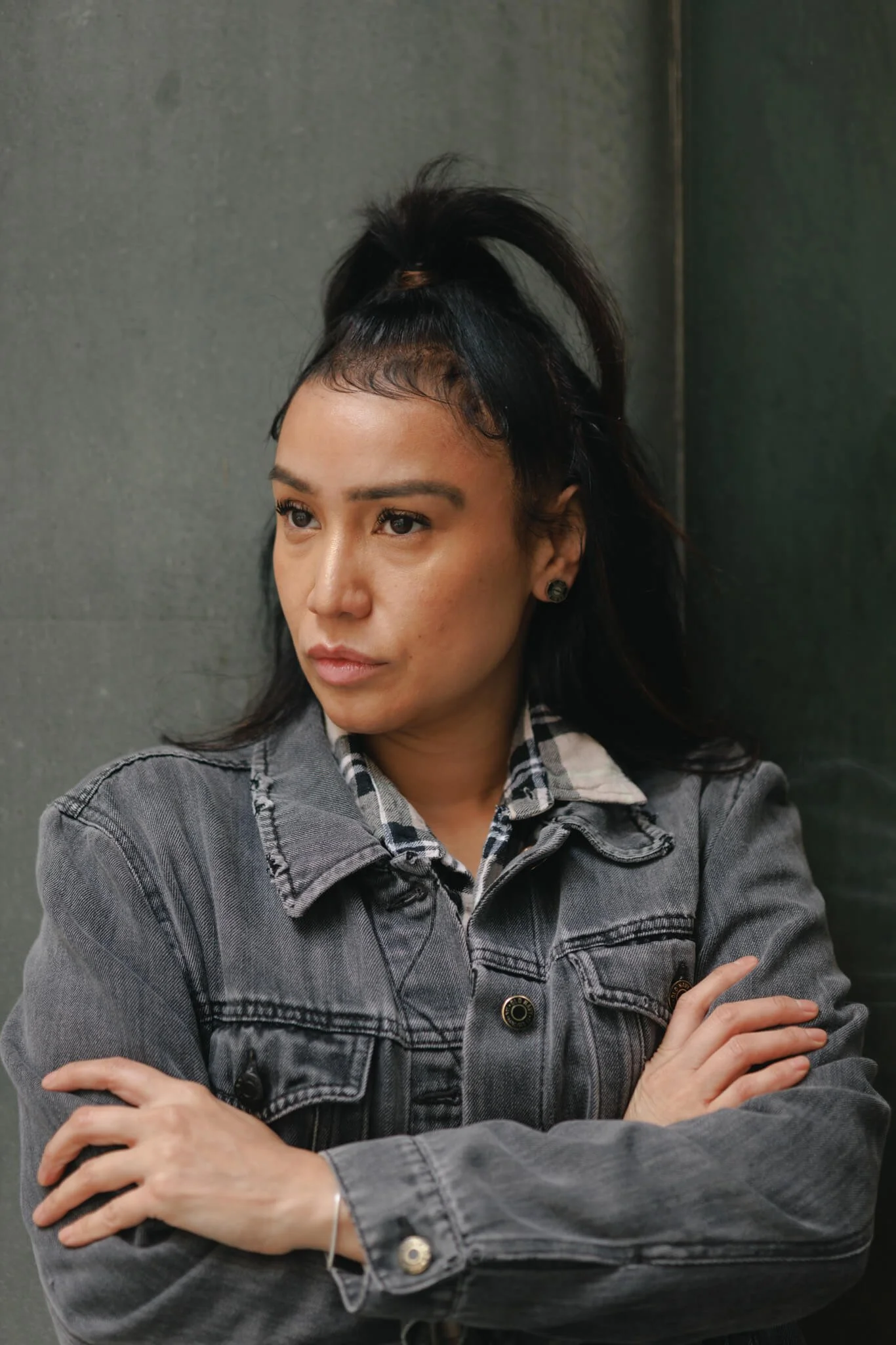 A woman with dark hair in a high ponytail with a brown hair tie, crossing her arms, wearing a gray denim jacket over a plaid shirt, in front of a dark gray wall.