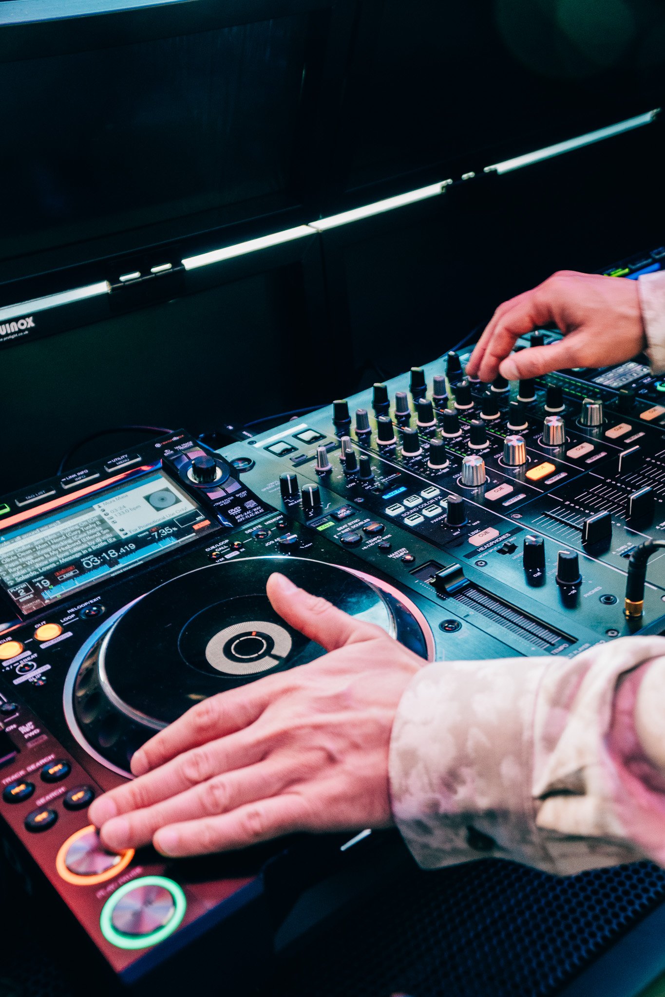 Close-up of DJ hands adjusting controls on a professional DJ mixer and CDJ turntable in a dark environment.