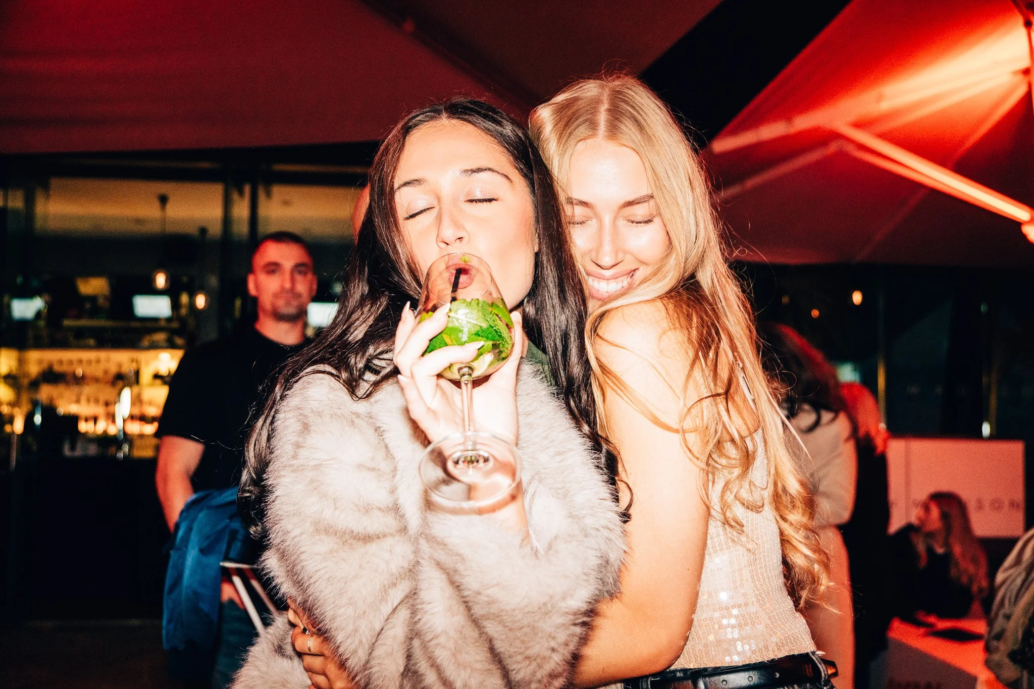 Two women with closed eyes smiling, one drinking a cocktail with mint and lime, the other leaning in close, at a party with bar and bartender behind them.