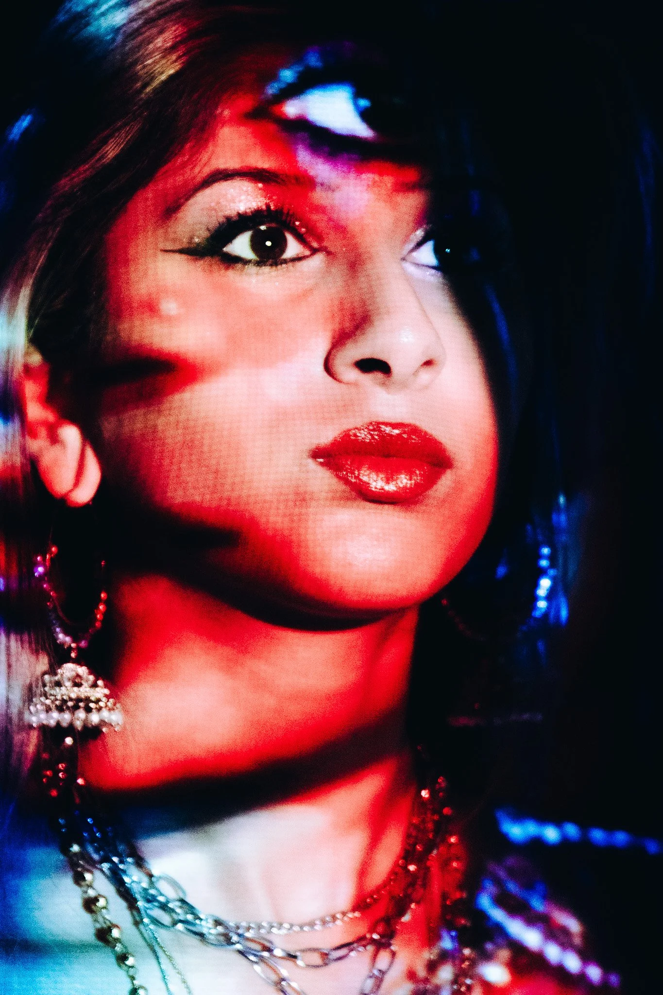 Close-up of a woman's face with makeup and jewelry, illuminated with colorful red, blue, and purple lights.