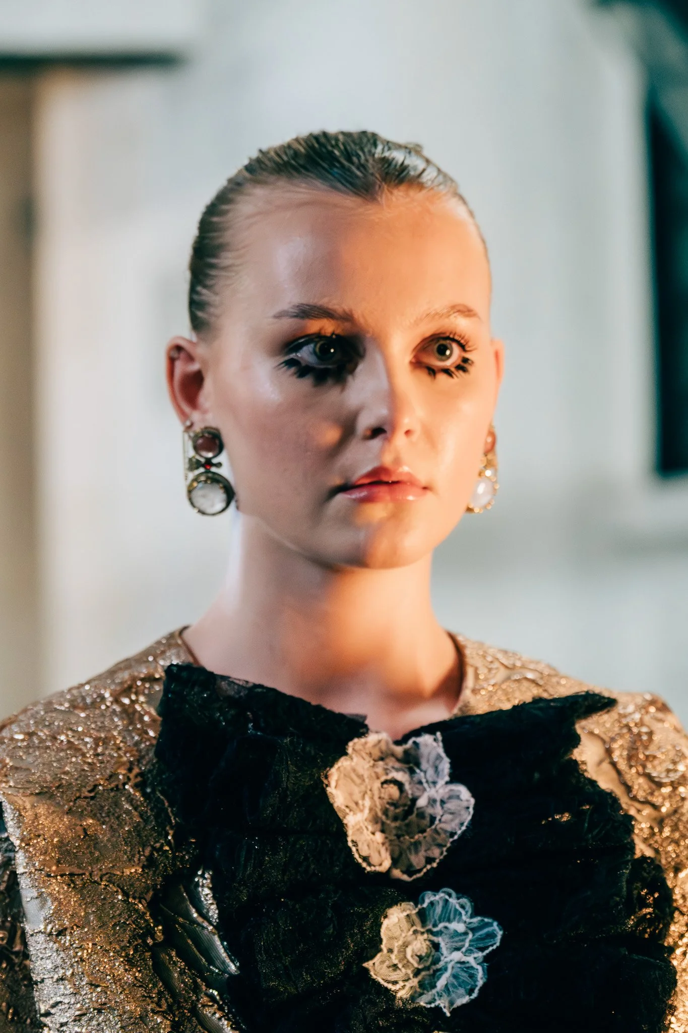 A woman with slicked-back hair and bold makeup, wearing large earrings and a gold and black embroidered dress with floral embellishments.