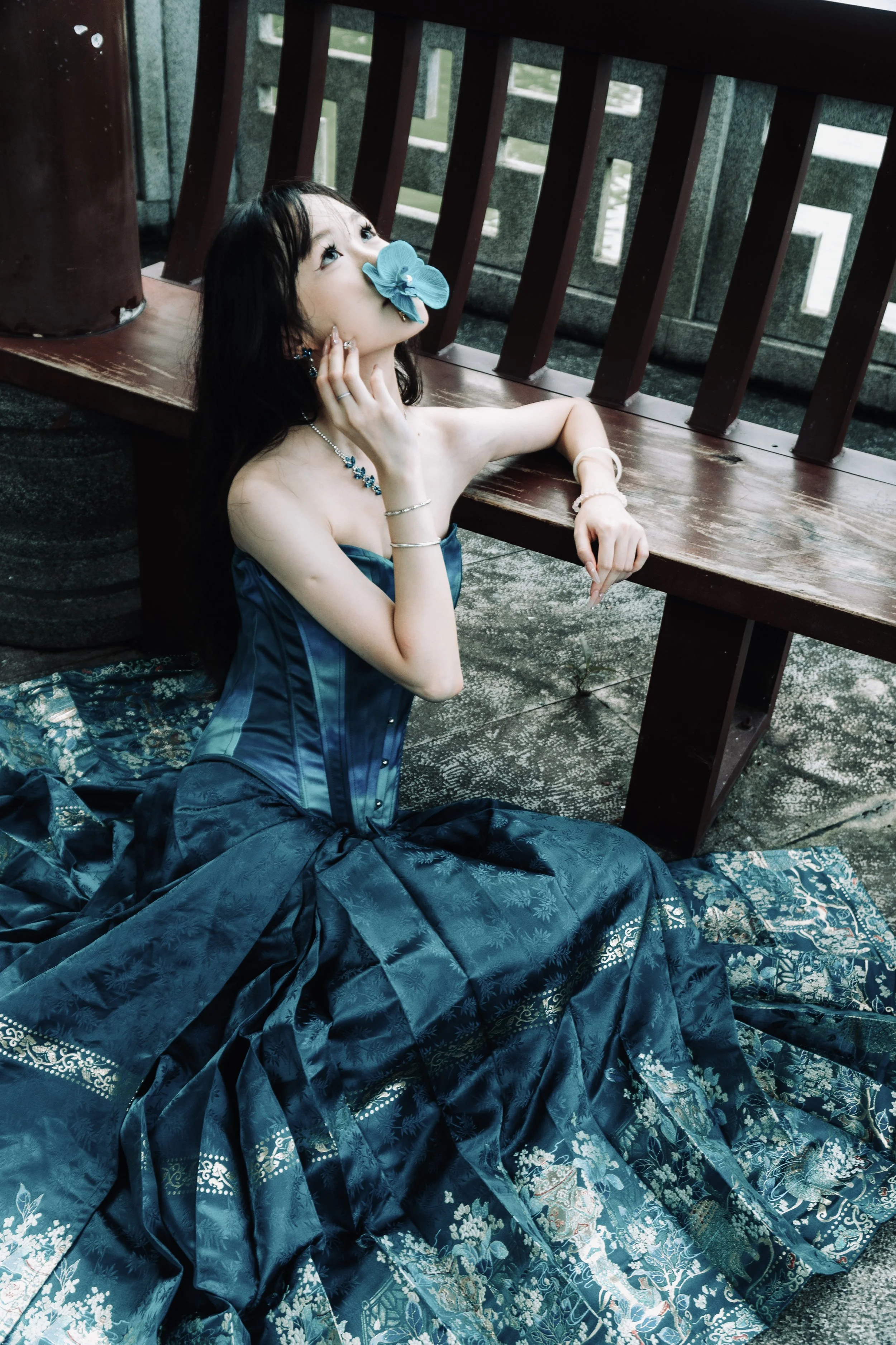 A woman in a blue dress with jewelry is seated on the ground, holding a blue flower to her nose, with a wooden bench and railing in the background.