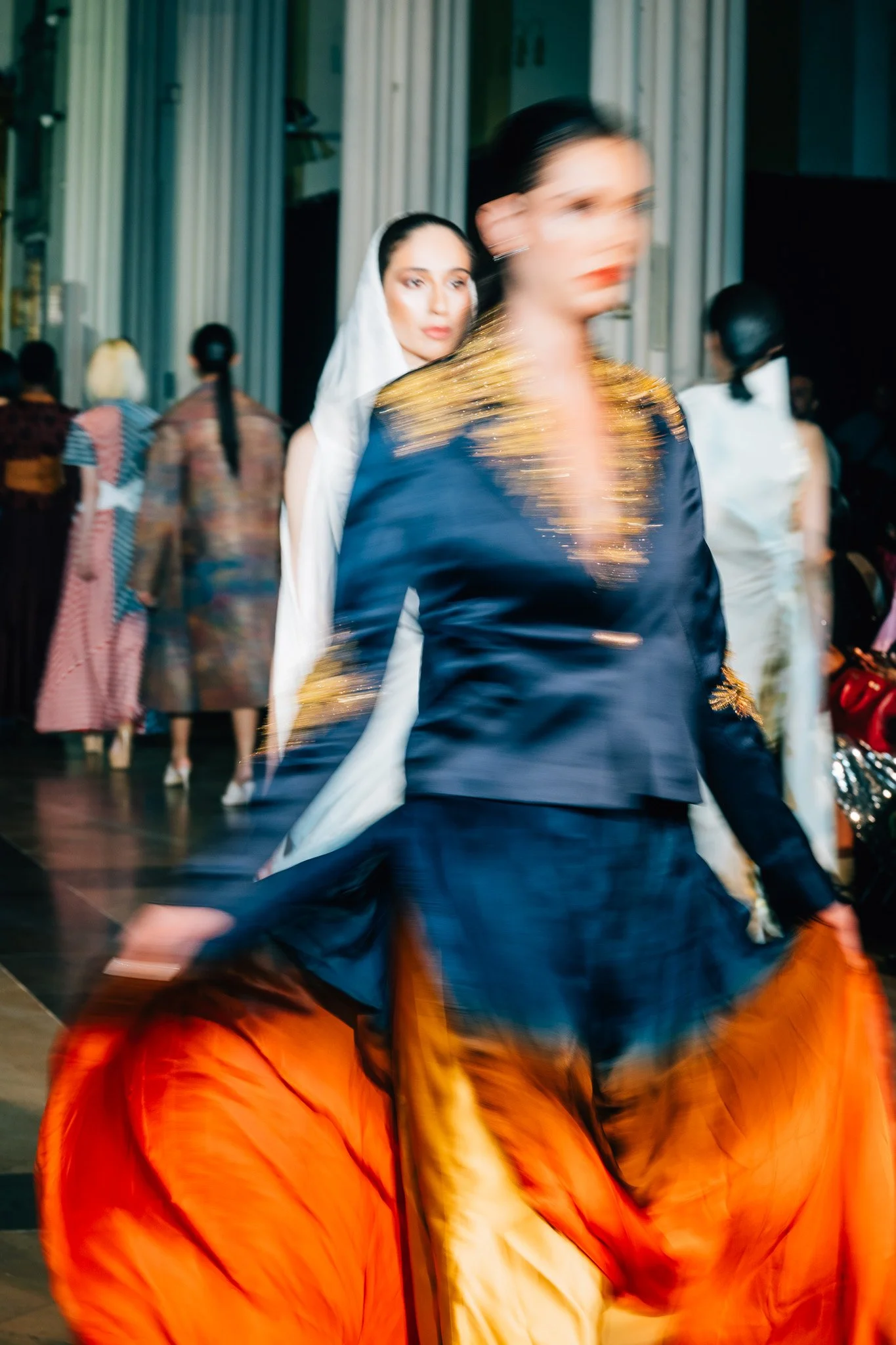 Blurred image of a fashion runway with models walking and wearing colorful outfits.