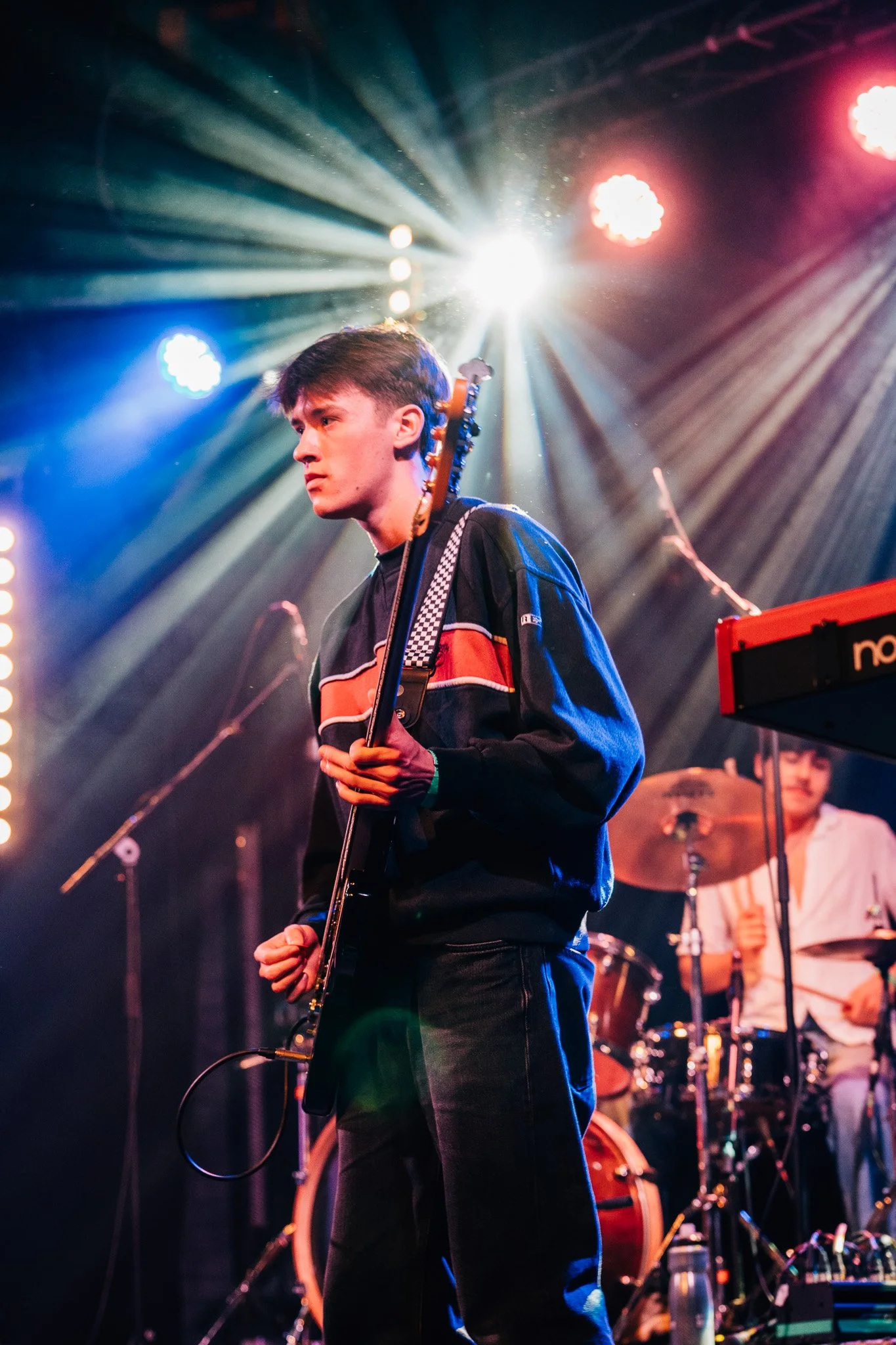 A young man playing an electric guitar on stage under multicolored lights, with a drummer in the background.
