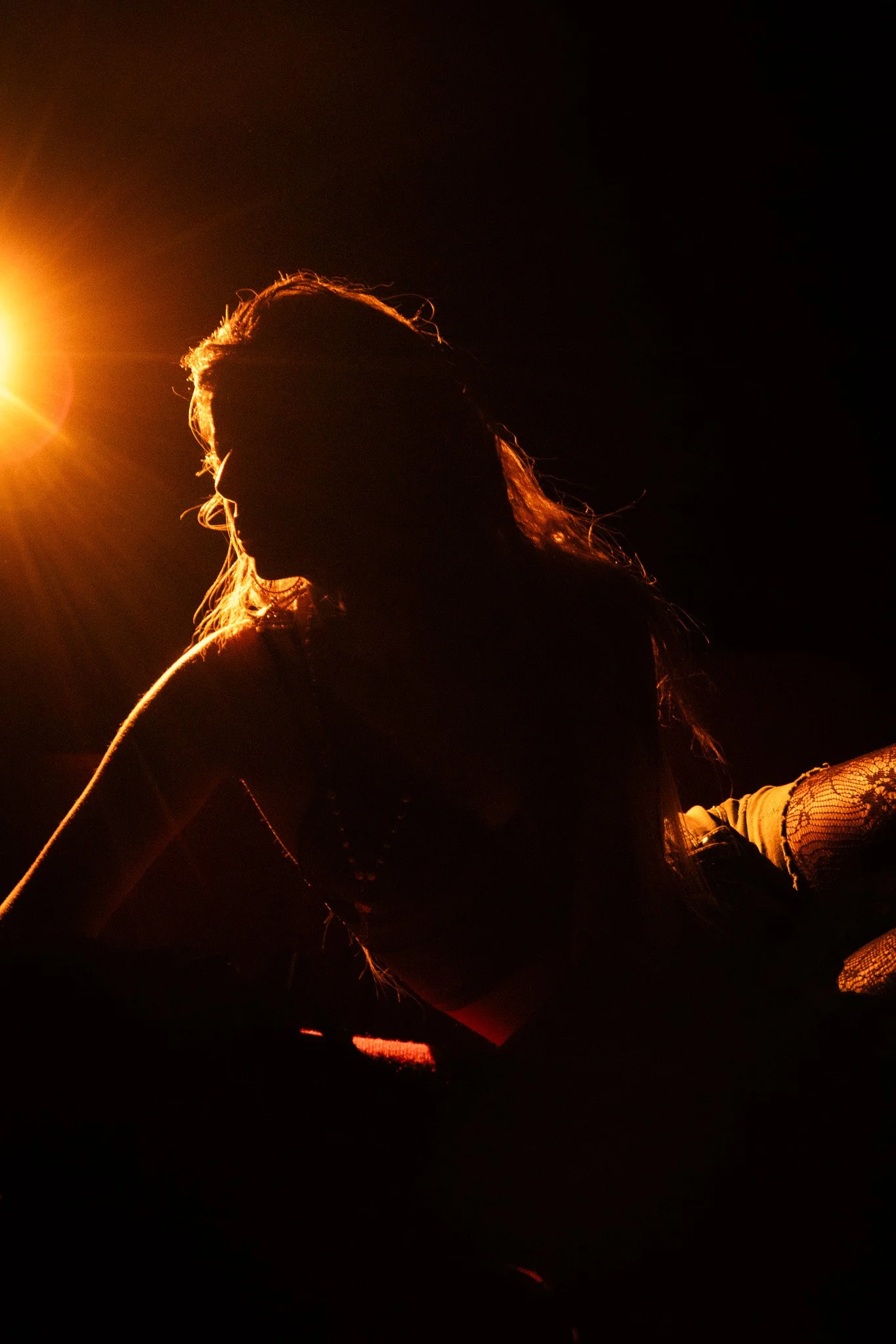 Silhouette of a woman with long hair illuminated by warm light, sitting with her head slightly bowed against a dark background.