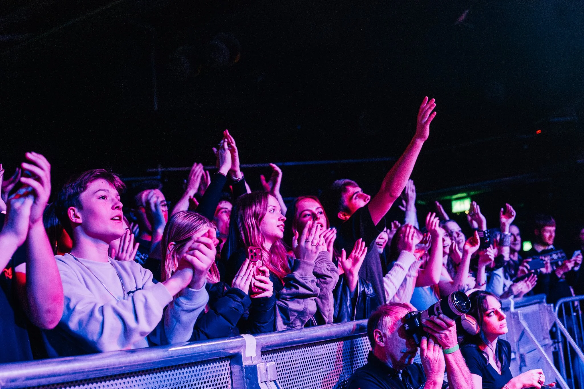 Audience at a concert or event, clapping and raising hands, with some holding cameras or smartphones, under vibrant stage lighting.