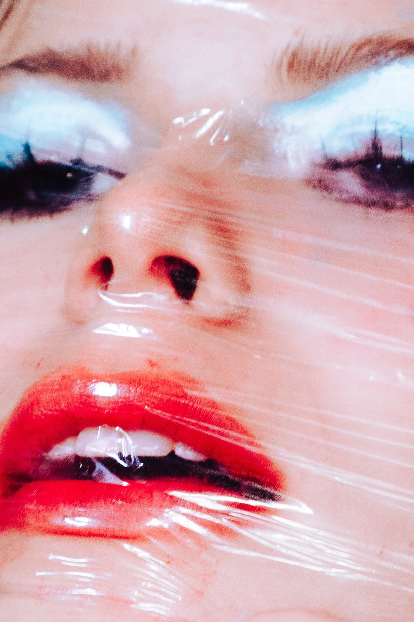 Close-up of lips painted in red lipstick with a reflection of a face and blue eye shadow, wrapped in transparent plastic.