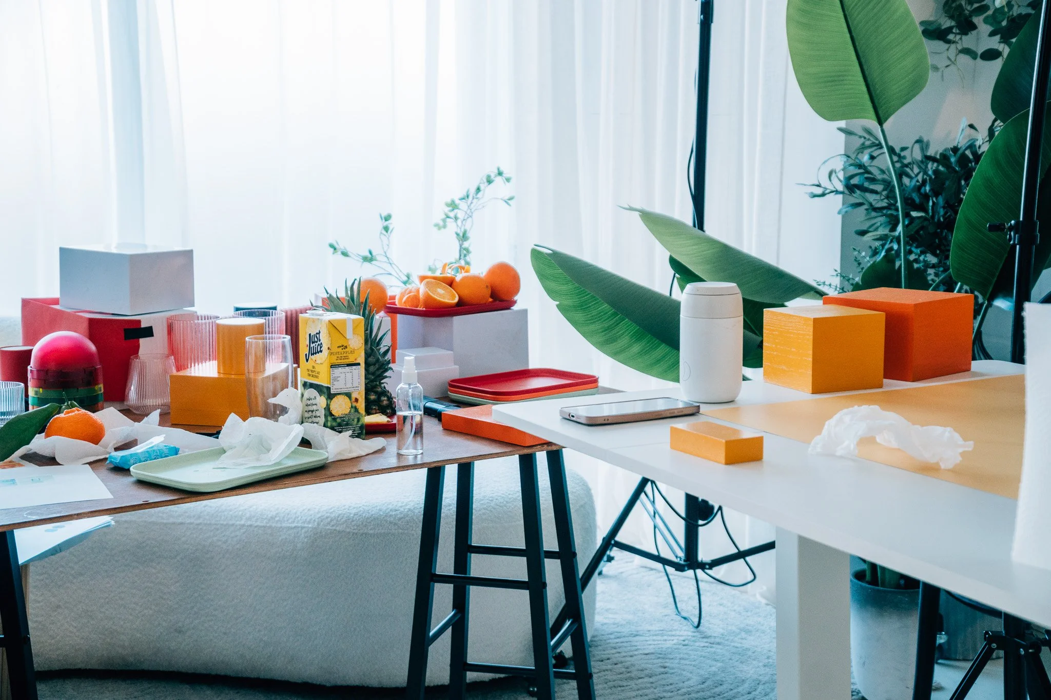 A cluttered workspace with various food and drink items, including oranges, juice boxes, cups, and miscellaneous objects on tables near large plants and a window with curtains.