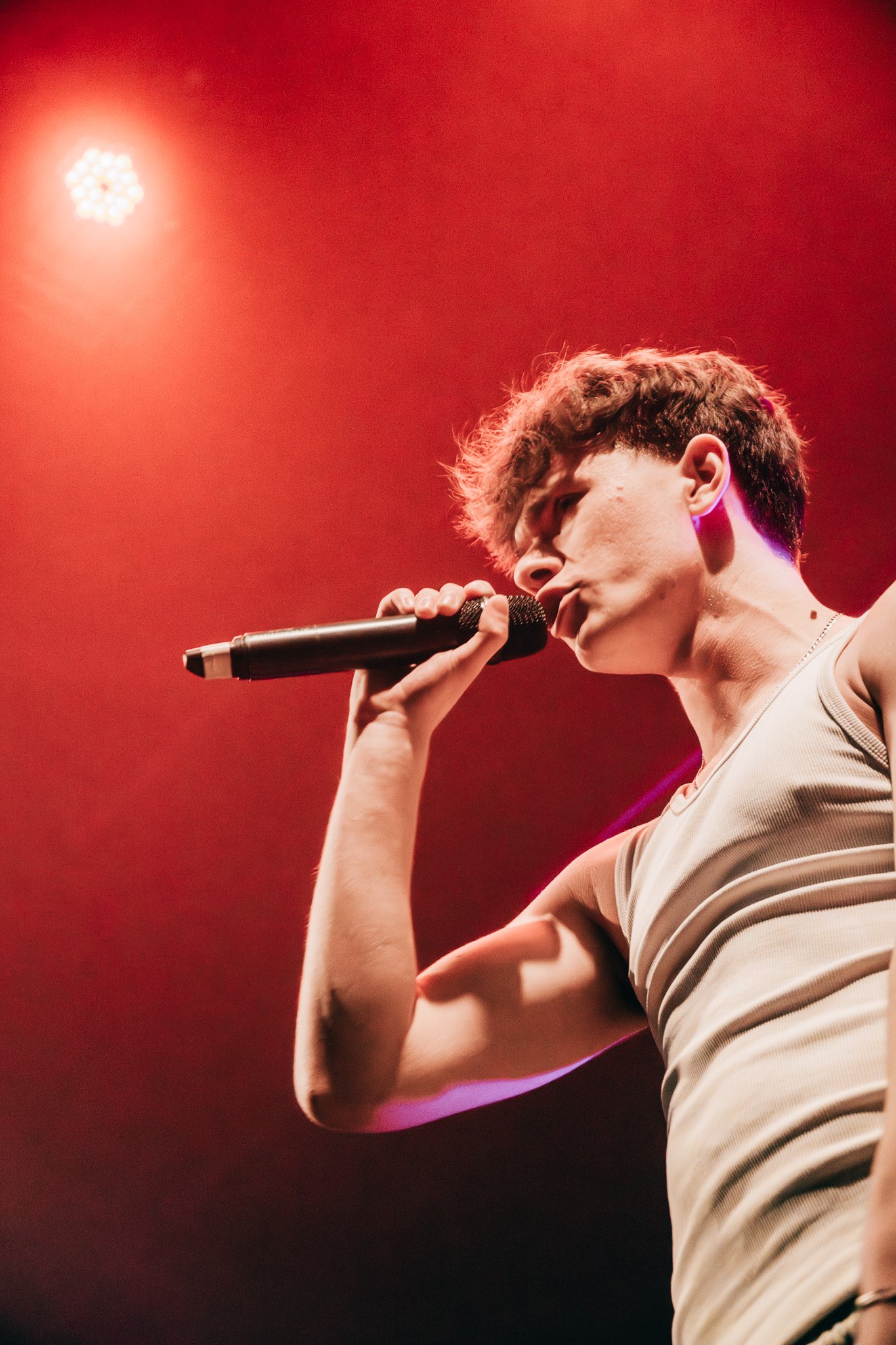 Young male singer performing on stage with red lighting, holding a microphone close to his mouth.