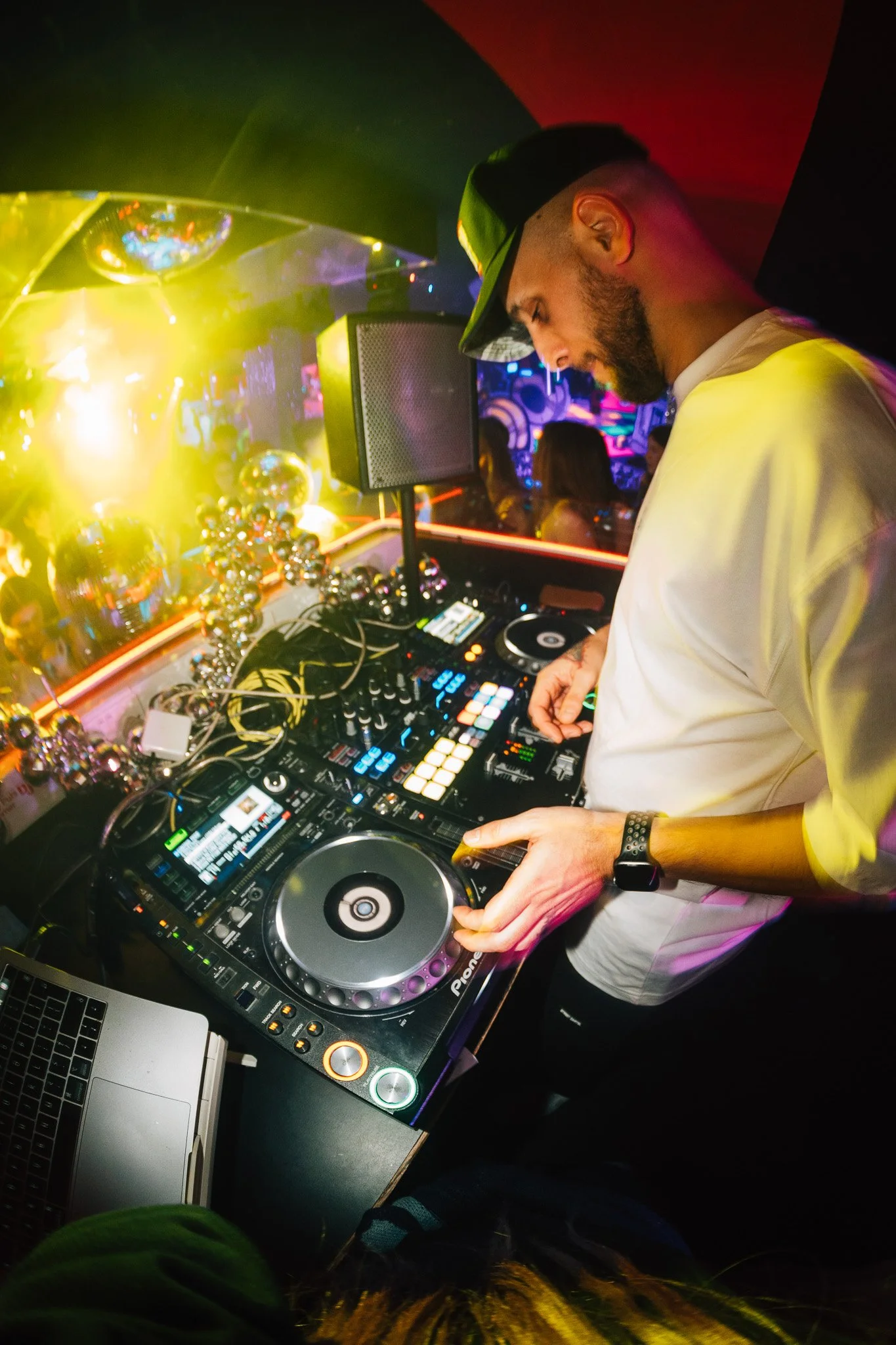 A male DJ wearing a white shirt and black cap, mixing music on DJ equipment at a nightclub with colorful lights and a crowd in the background.