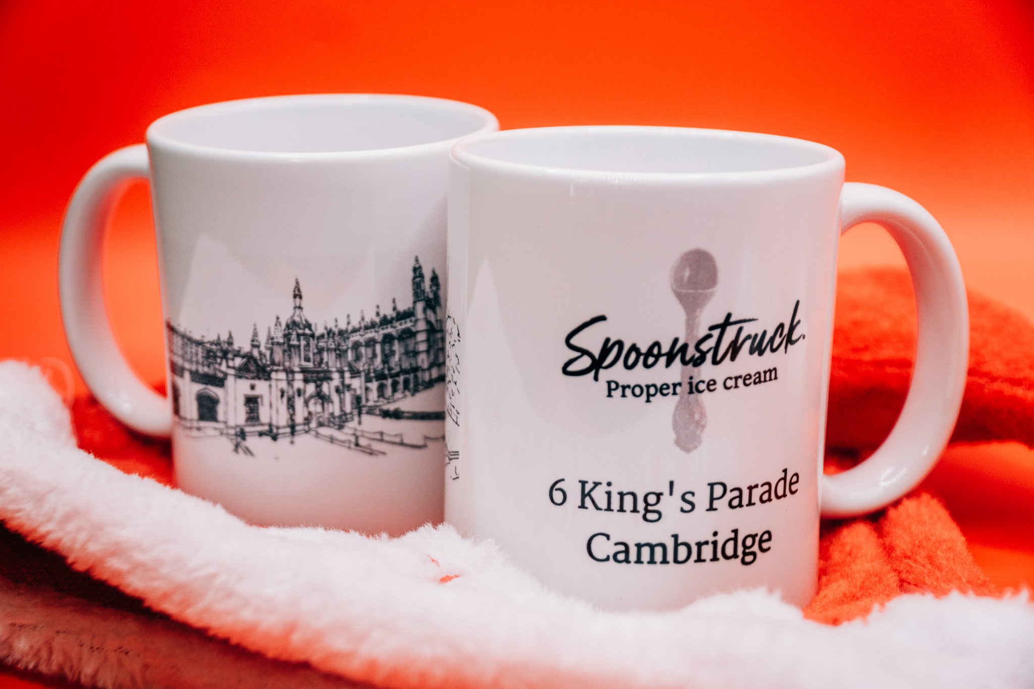 Close-up of two white coffee mugs with black print, showing a cityscape and the word 'Spoontrunk' on the mugs, set against a vibrant orange background.