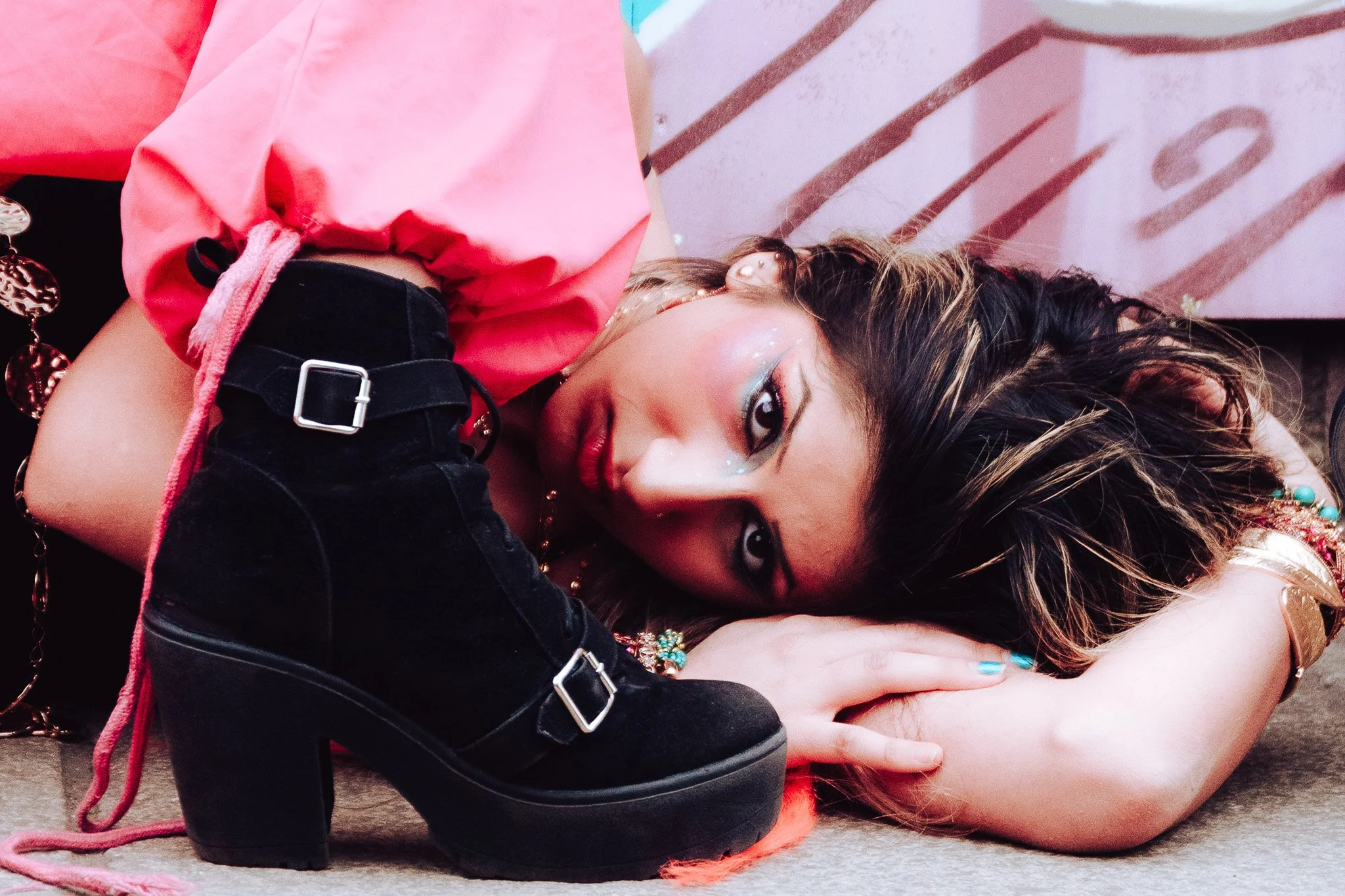 A woman lying on the ground with her head turned to the side, wearing colorful makeup and earrings, with her arm covering part of her face. She is dressed in a pink top and black pants with metal buckles, and there are jewelry accents nearby. The set