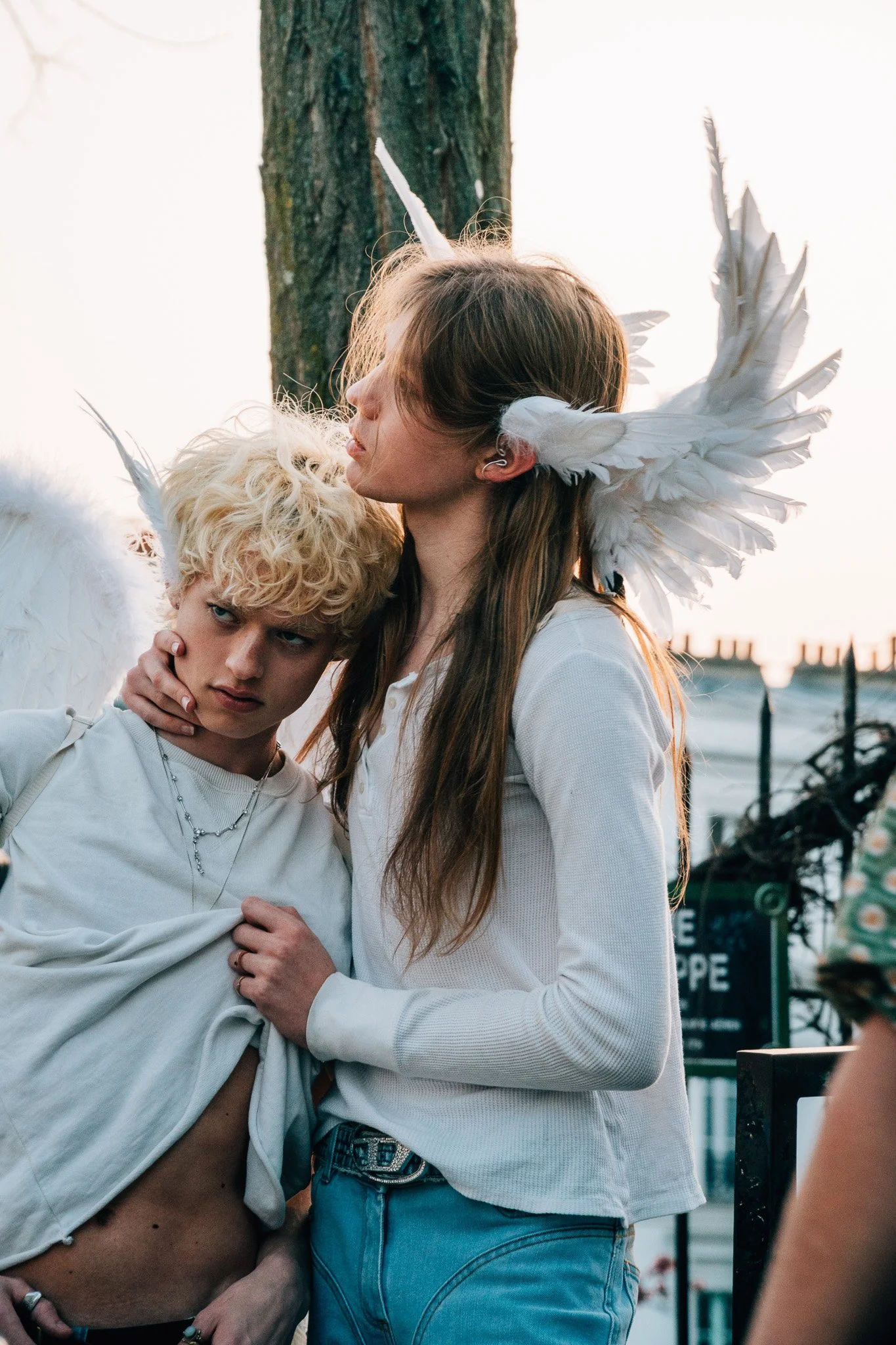 Two young women, one with angel wings and the other with devil horns, embracing outdoors at sunset.