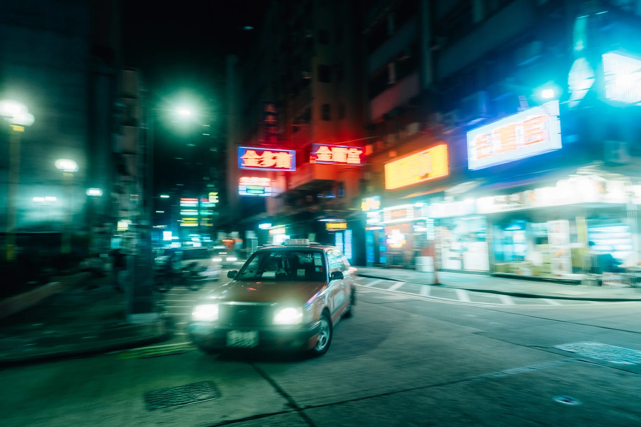 A city street at night with neon signs and blurred lights, including a taxi, cars, and buildings.